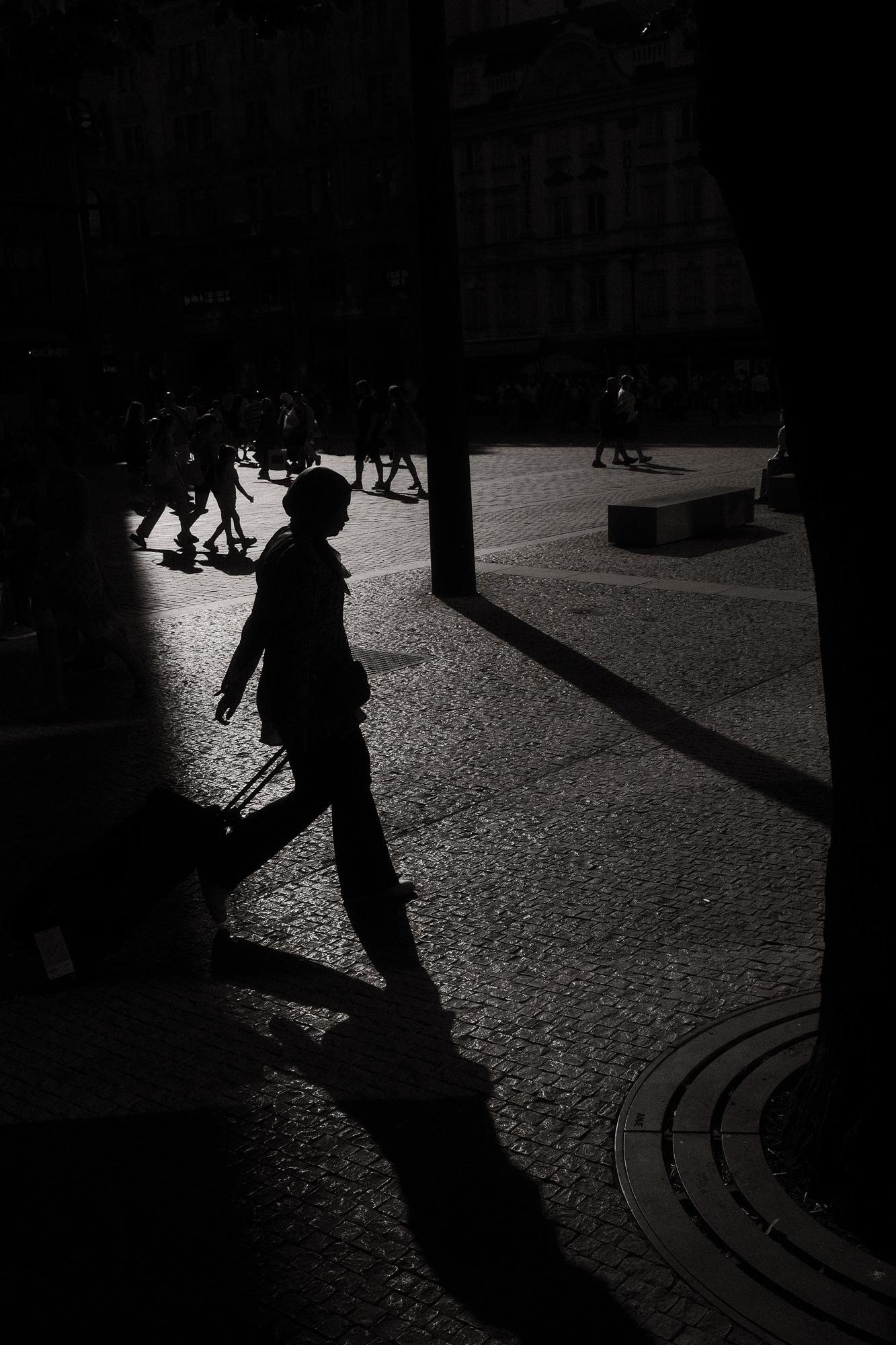 Silueta ženy s cestovnou taškou na kolieskach, ktorá kráča po dláždenom námestí v protisvetle.