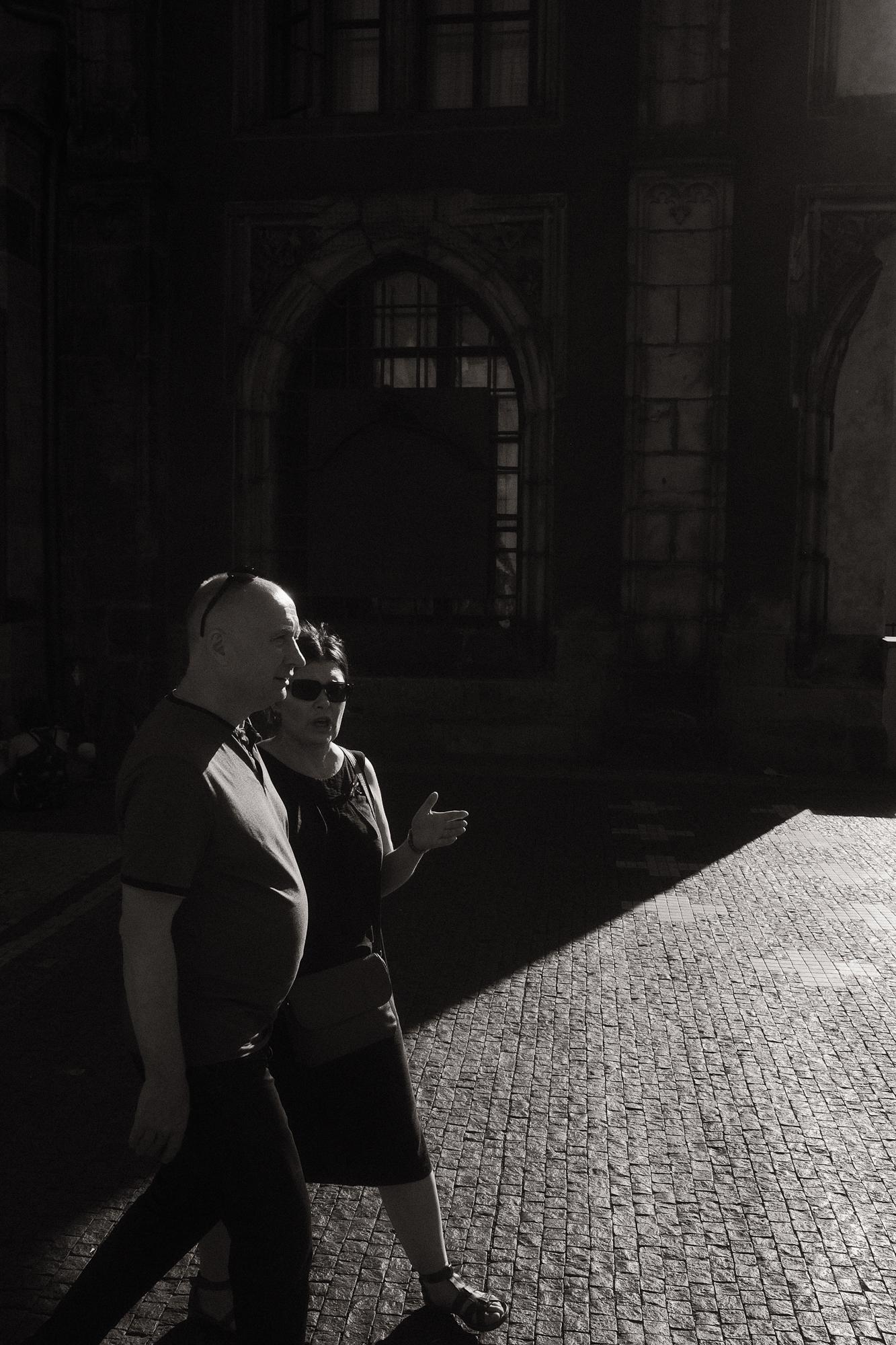 Čiernobiela fotografia zachytáva dvoch ľudí, ktorí kráčajú po dláždenom námestí v tieni historickej budovy.