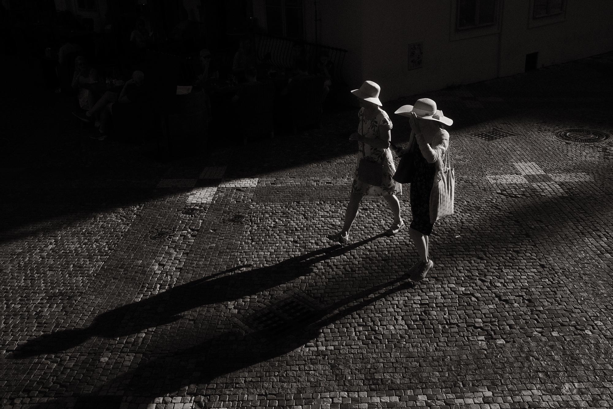 Dve ženy v klobúkoch kráčajú po kamennej ulici, osvetlené šikmým slnečným svetlom, s dlhými tieňmi na zemi.