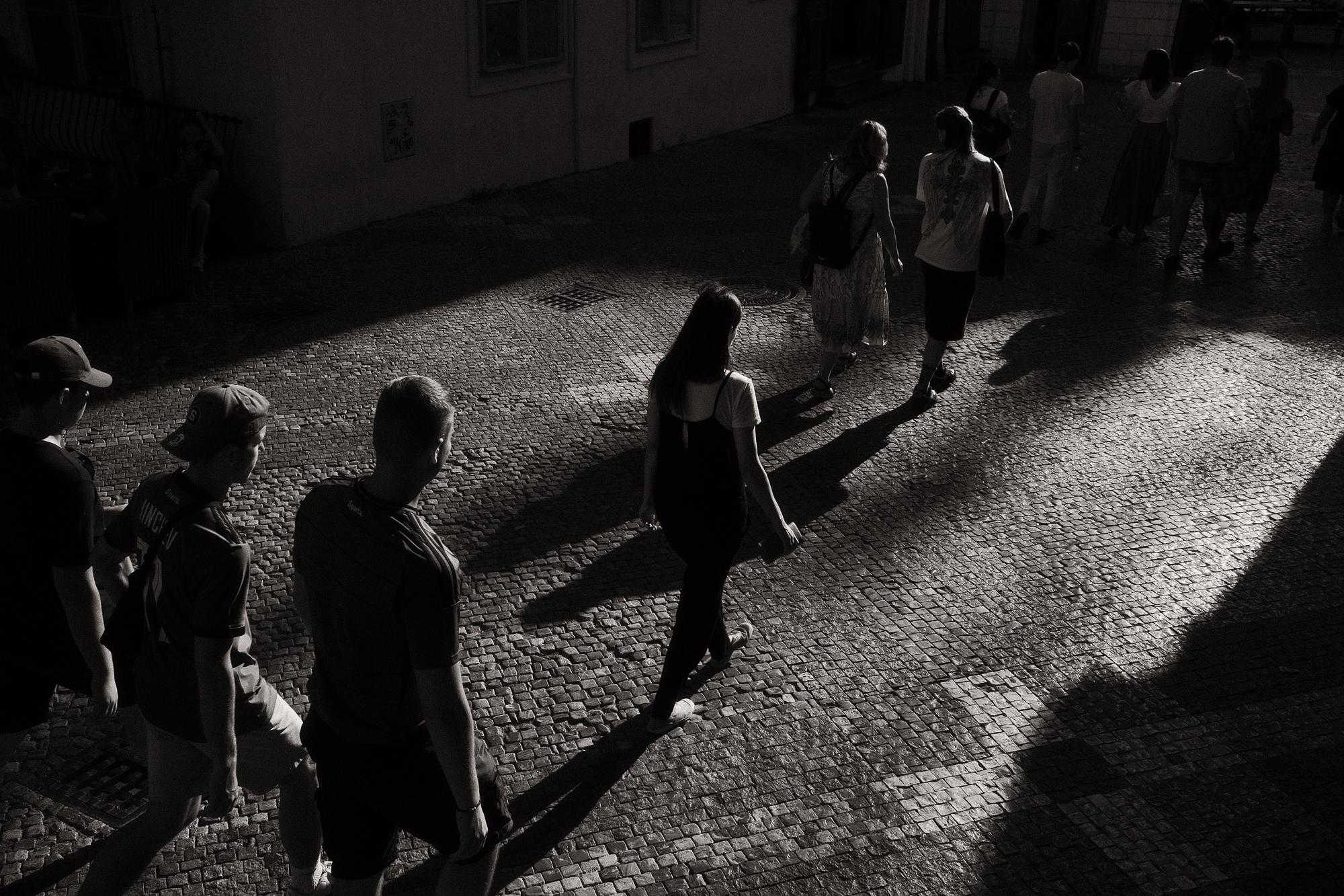 Ulica so skupinou ľudí kráčajúcich cez dlažbu, osvetlenú dramatickými tieňmi, vytvára čiernobiely kontrast.