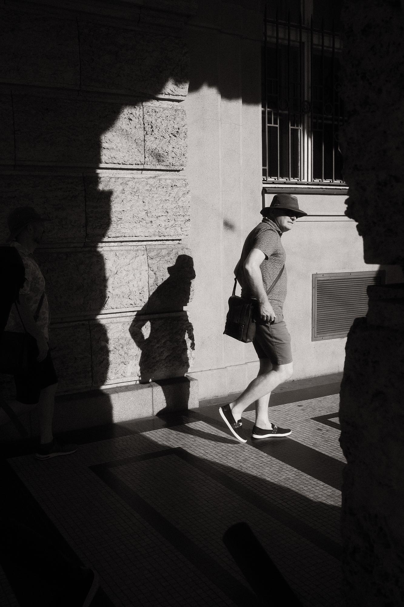 Čiernobiela fotografia muža vchádzajúceho do slnka, jeho ostrý tieň je vrhaný na textúrovanú kamennú stenu.