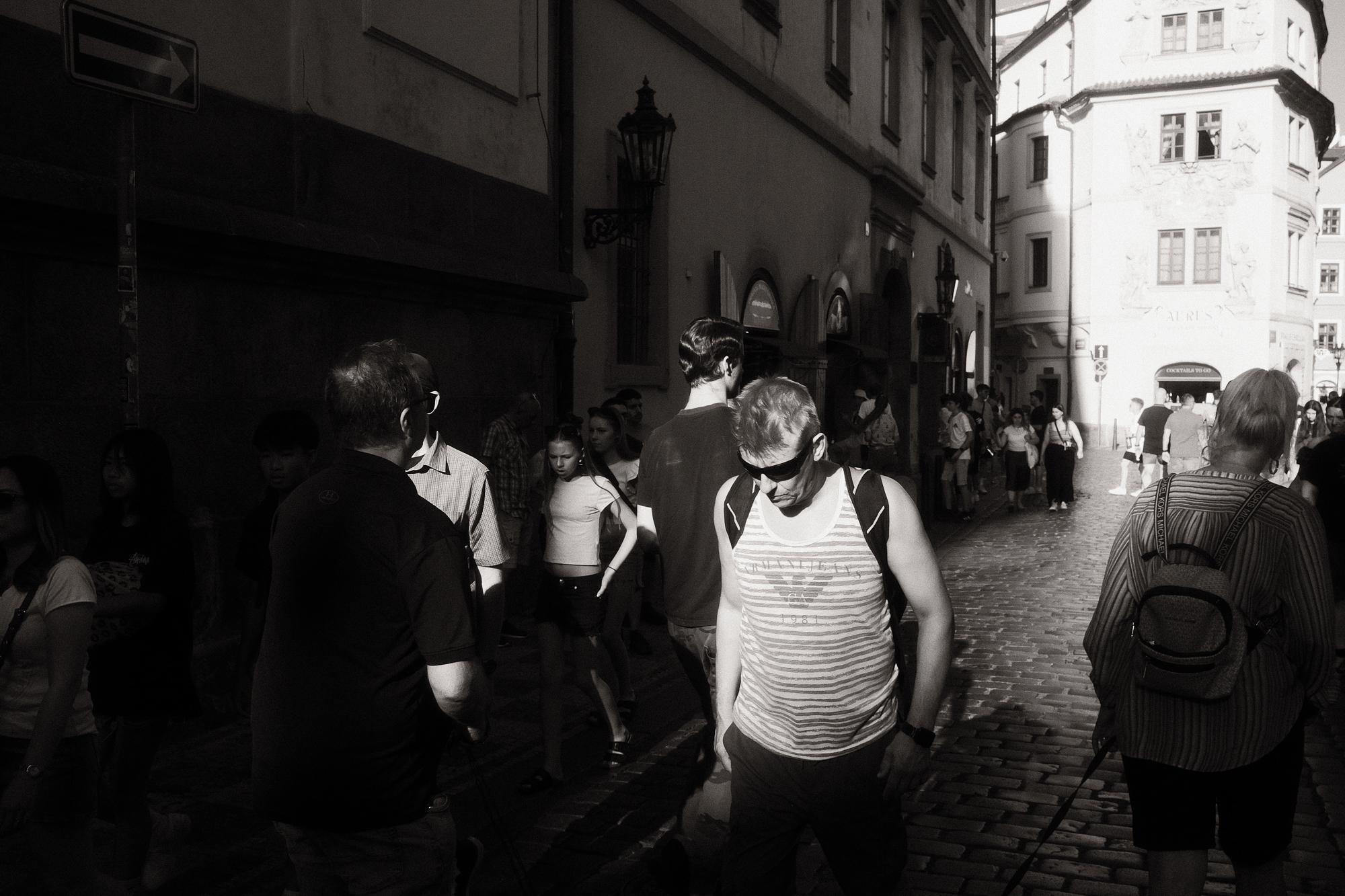 Čiernobiela fotografia zobrazujúca frekventovanú uličku, kde sa ľudia pohybujú po dláždenej dlažbe.