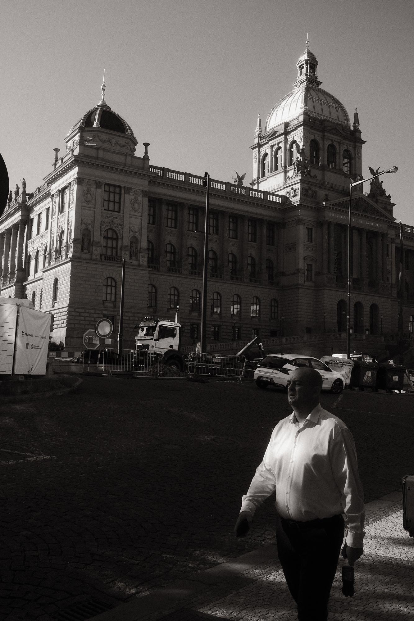 Čiernobiela fotografia muža v bielej košeli kráčajúceho popri historickej budove s majestátnou kopulou v pozadí.