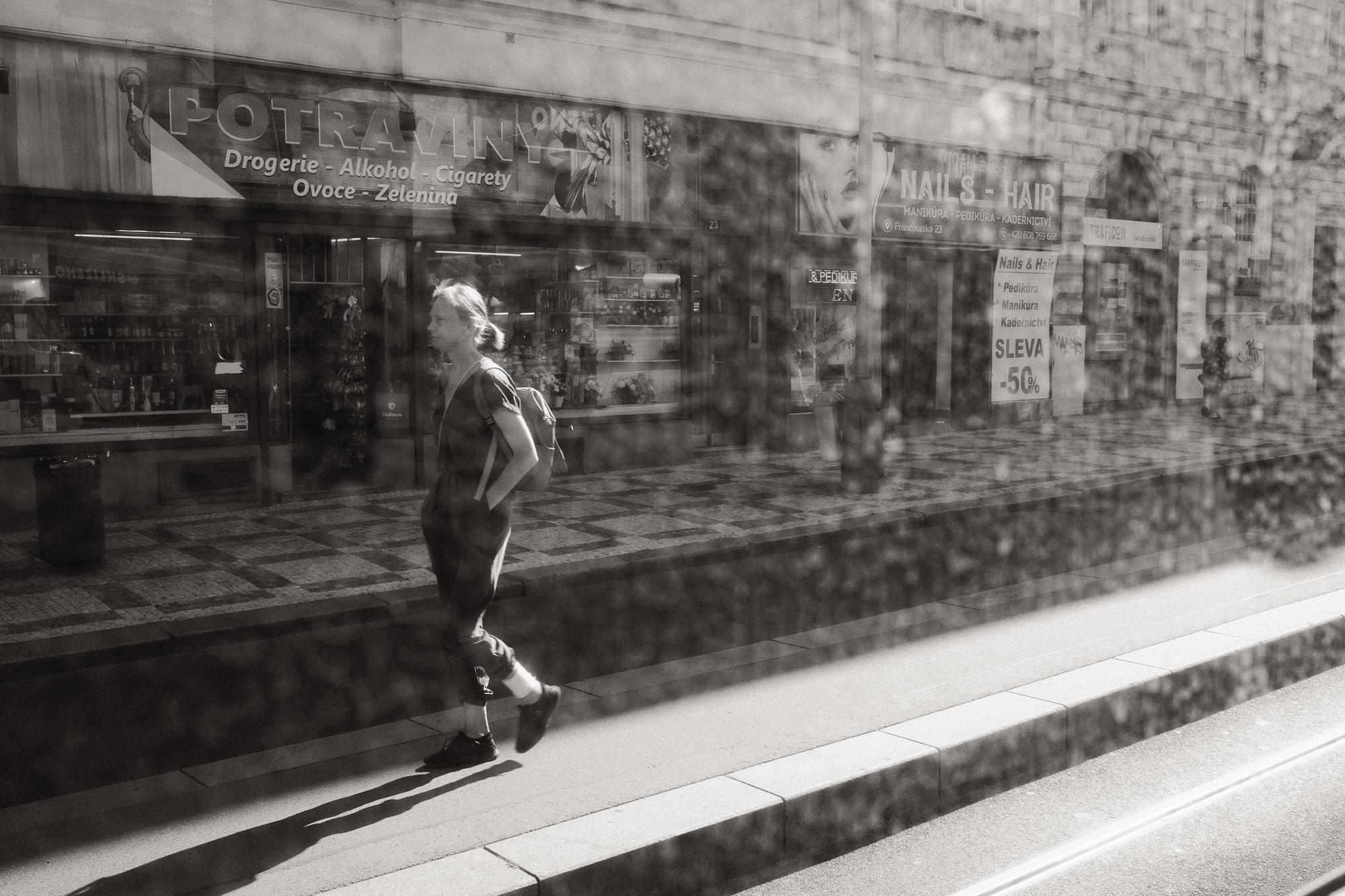 Čiernobiela fotografia ženy kráčajúcej po chodníku pred obchodmi, fotografované cez špinavé okno električky.
