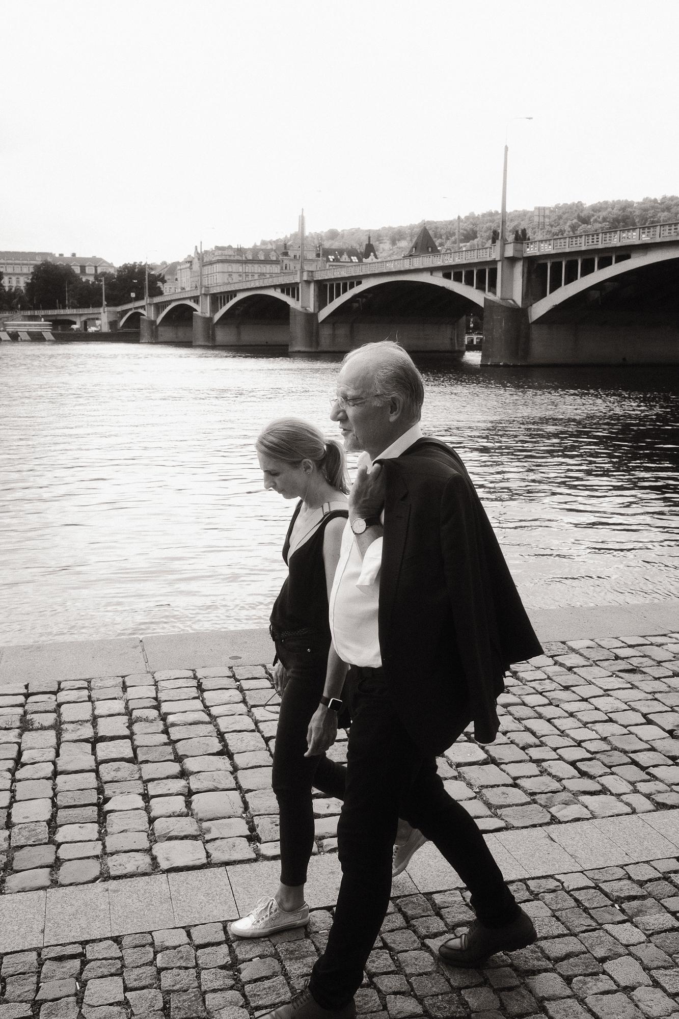 Čiernobiela fotografia dvoch osôb kráčajúcich popri rieke s kamenným mostom v pozadí.