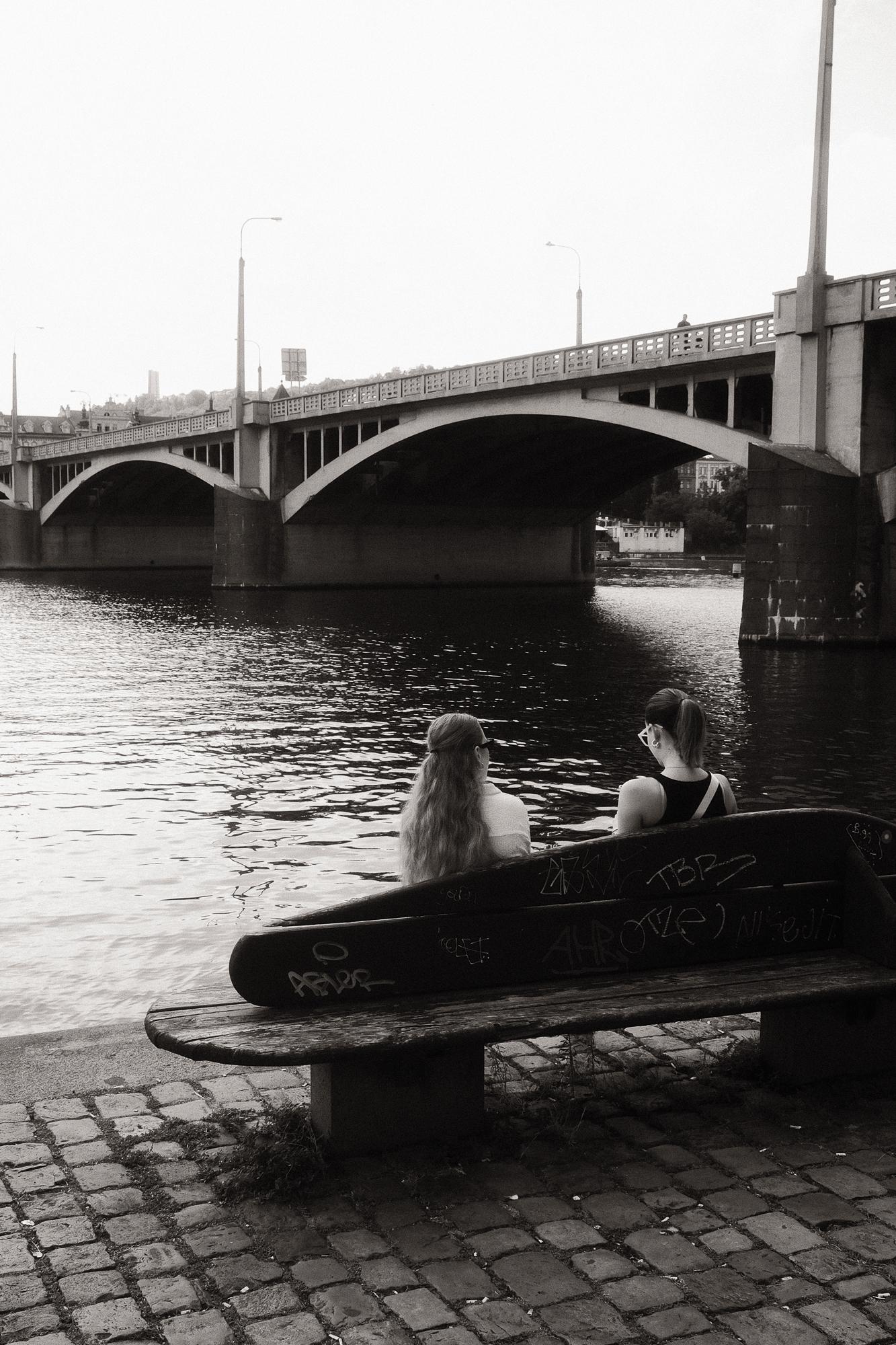 Dve ženy sedia na lavici pri Vltave na náplavke.