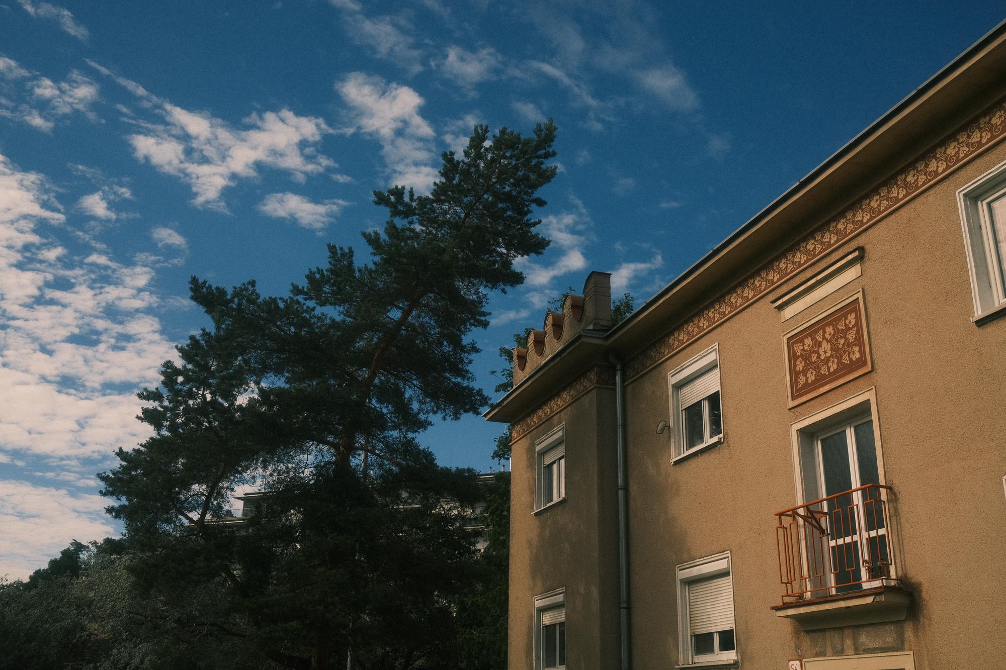 Ulica s historickou budovou s ornamentálnymi detailmi a zeleňou na pozadí pod modrou oblohou s oblakmi. Pezinok