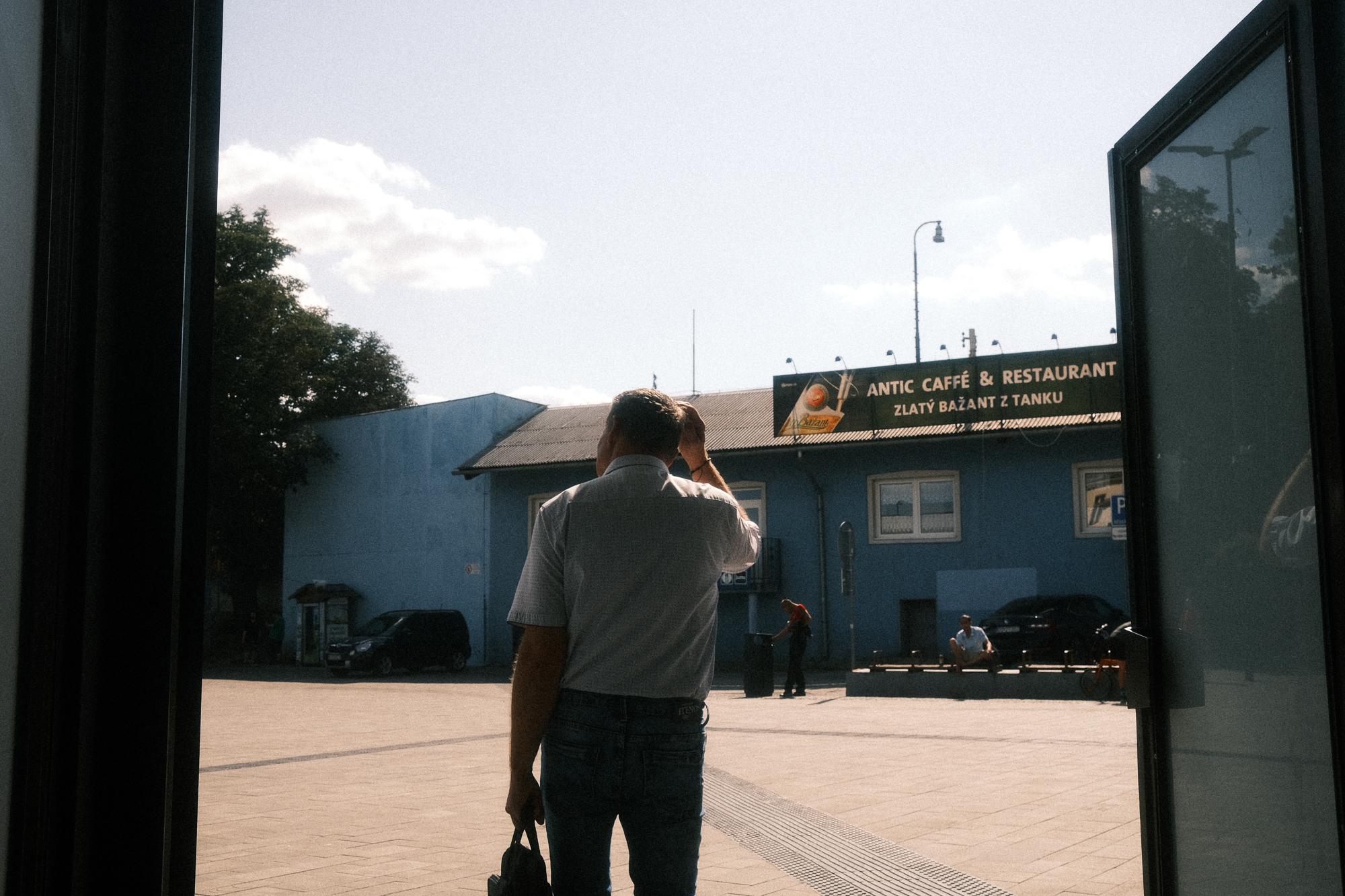 Muž vychádzajúci do slnečného dňa z Bratislavskej hlavnej stanice orámovaný vchodovými dverami.