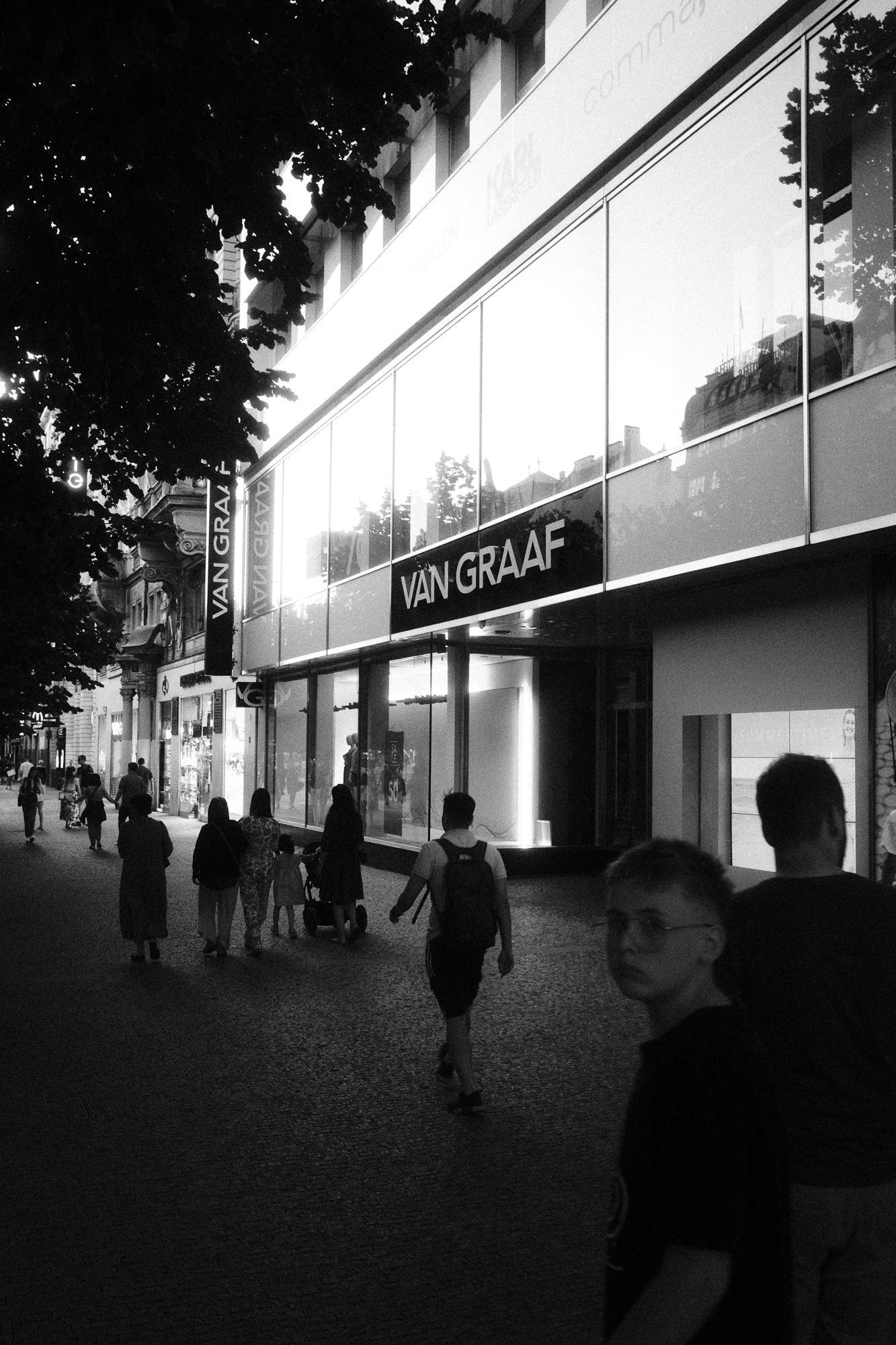 Čiernobiela fotografia ulice so skupinou ľudí kráčajúcich po chodníku vedľa moderných obchodov. Jeden chlapec pozerá priamo do objektívu.