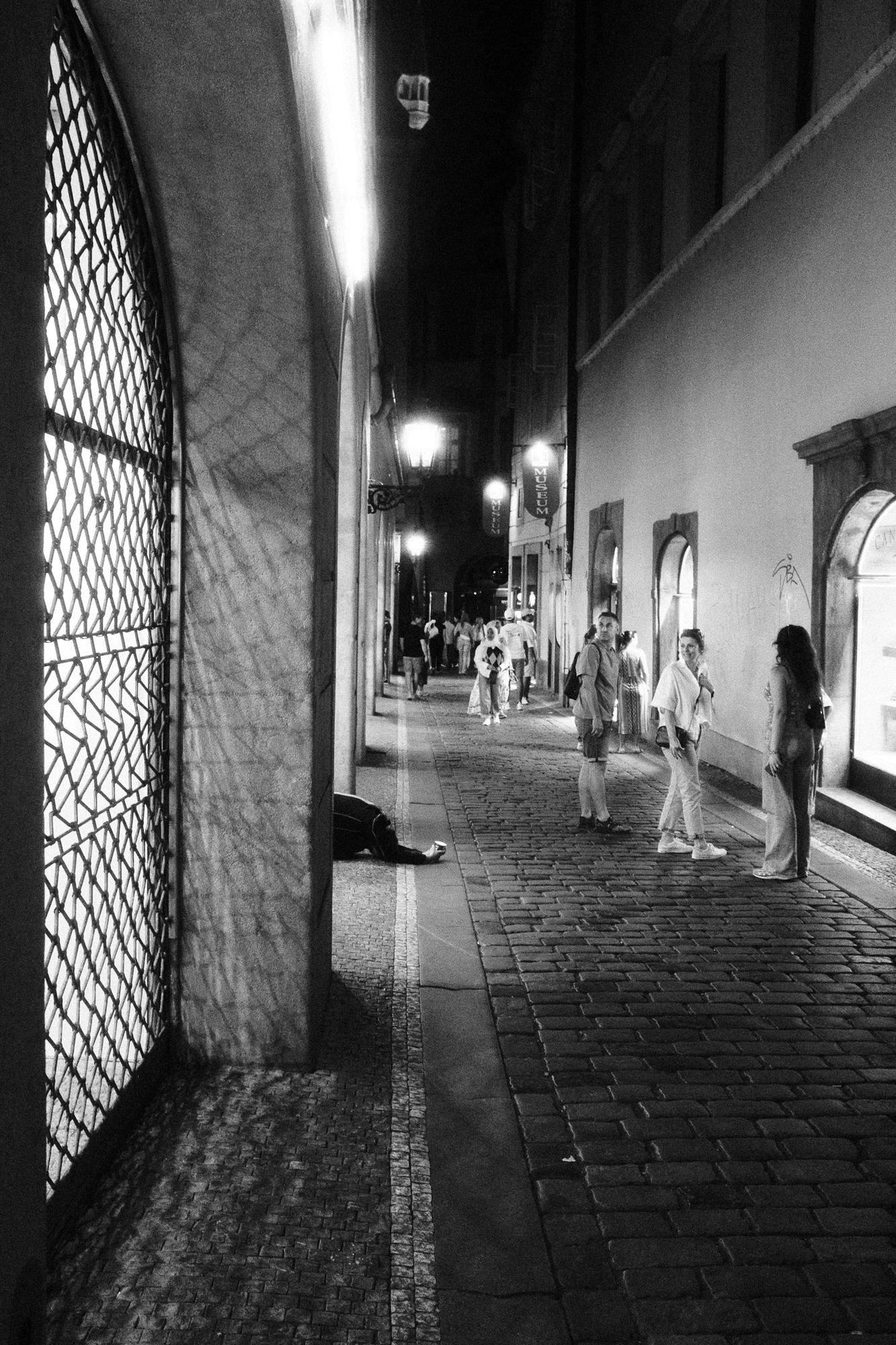 Čiernobiela fotografia zobrazujúca ľudí stojacich oproti žobrákovi v rušnej nočnej ulici.