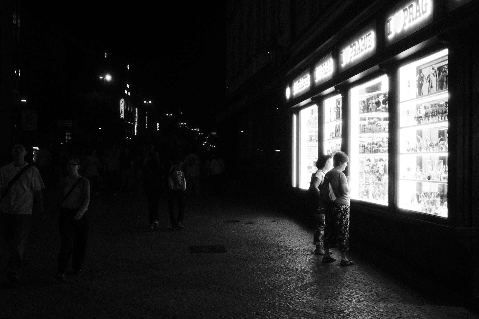 Čiernobiela fotografia ľudí prechádzajúcich sa nočnou ulicou. Dve ženy si prezerajú tovar osvetlené svetlom z výkladu.