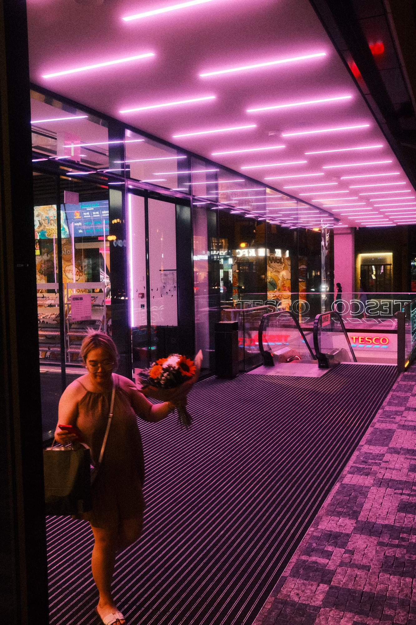 Žena kráča z moderného obchodu s kvetmi a telefónom v ruke pod fialovým neonovým osvetlením.