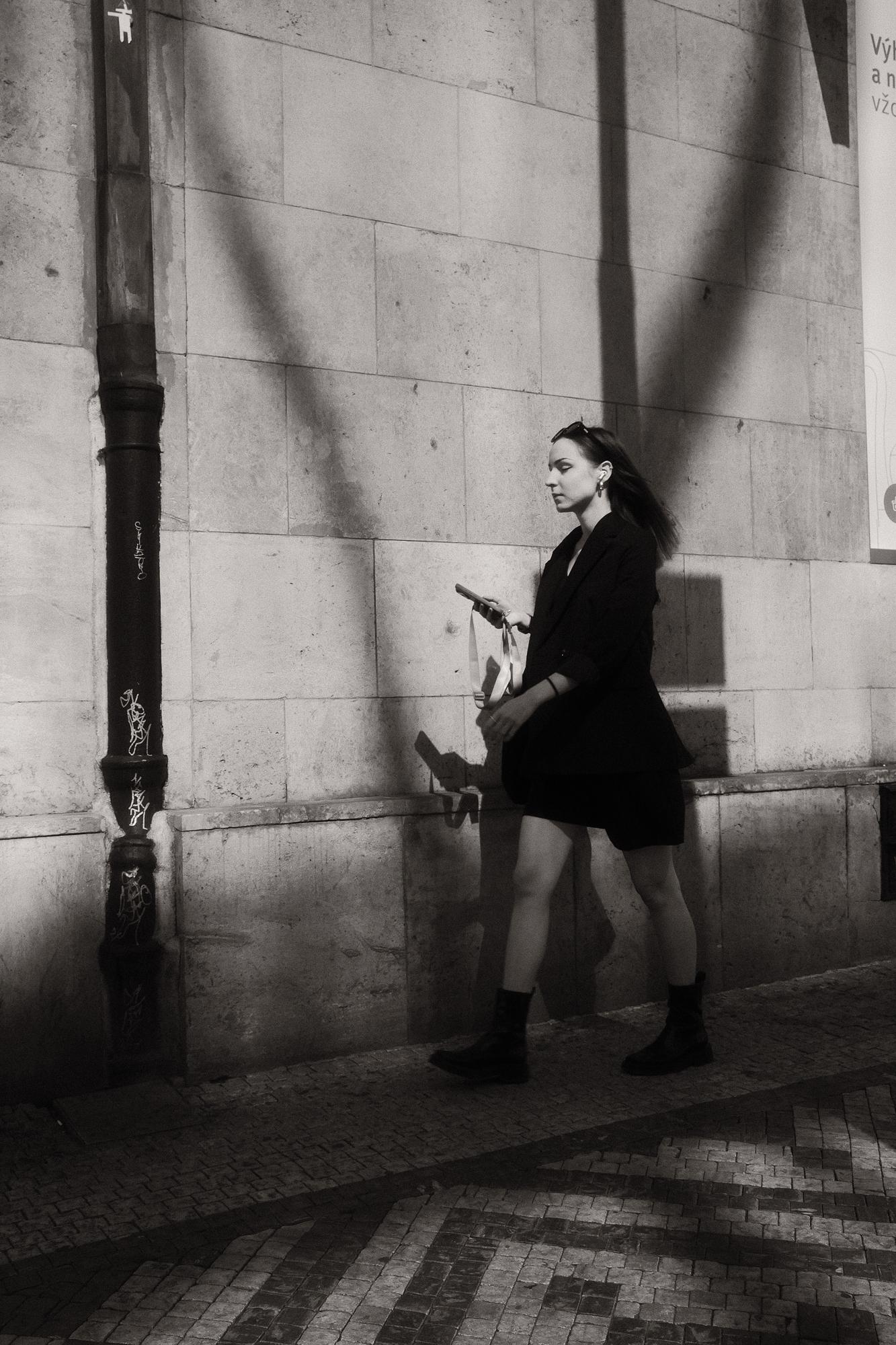 Čiernobiela fotografia ženy kráčajúcej popri stene v meste s mobilom v ruke. Slnečné lúče vytvárajú tiene na stene a zemi.