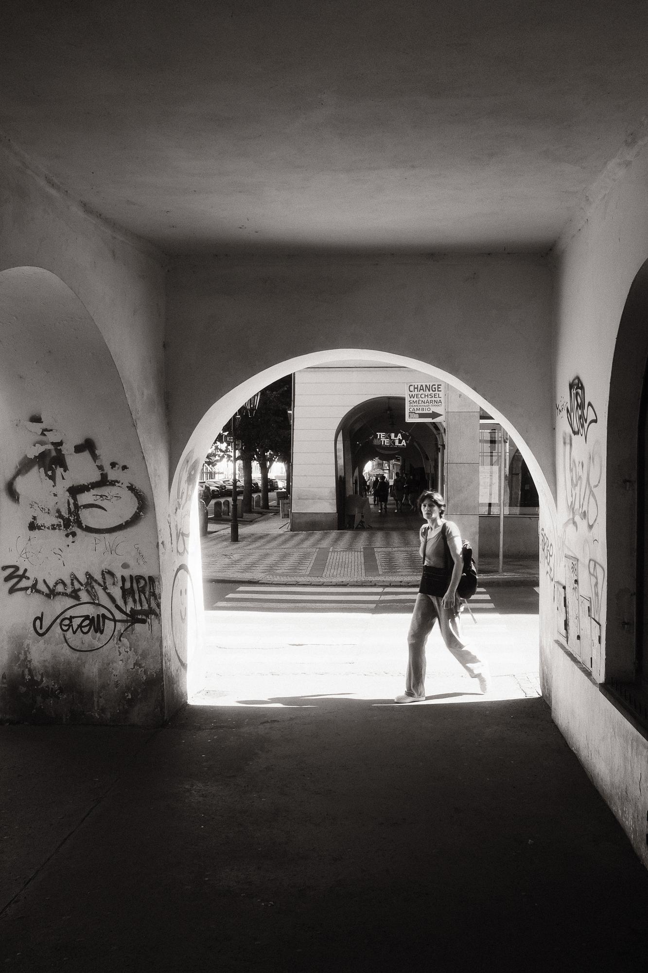 Čiernobiela fotografia osoby kráčajúcej cez osvetlený oblúk prechodu, ozdobeného graffiti.