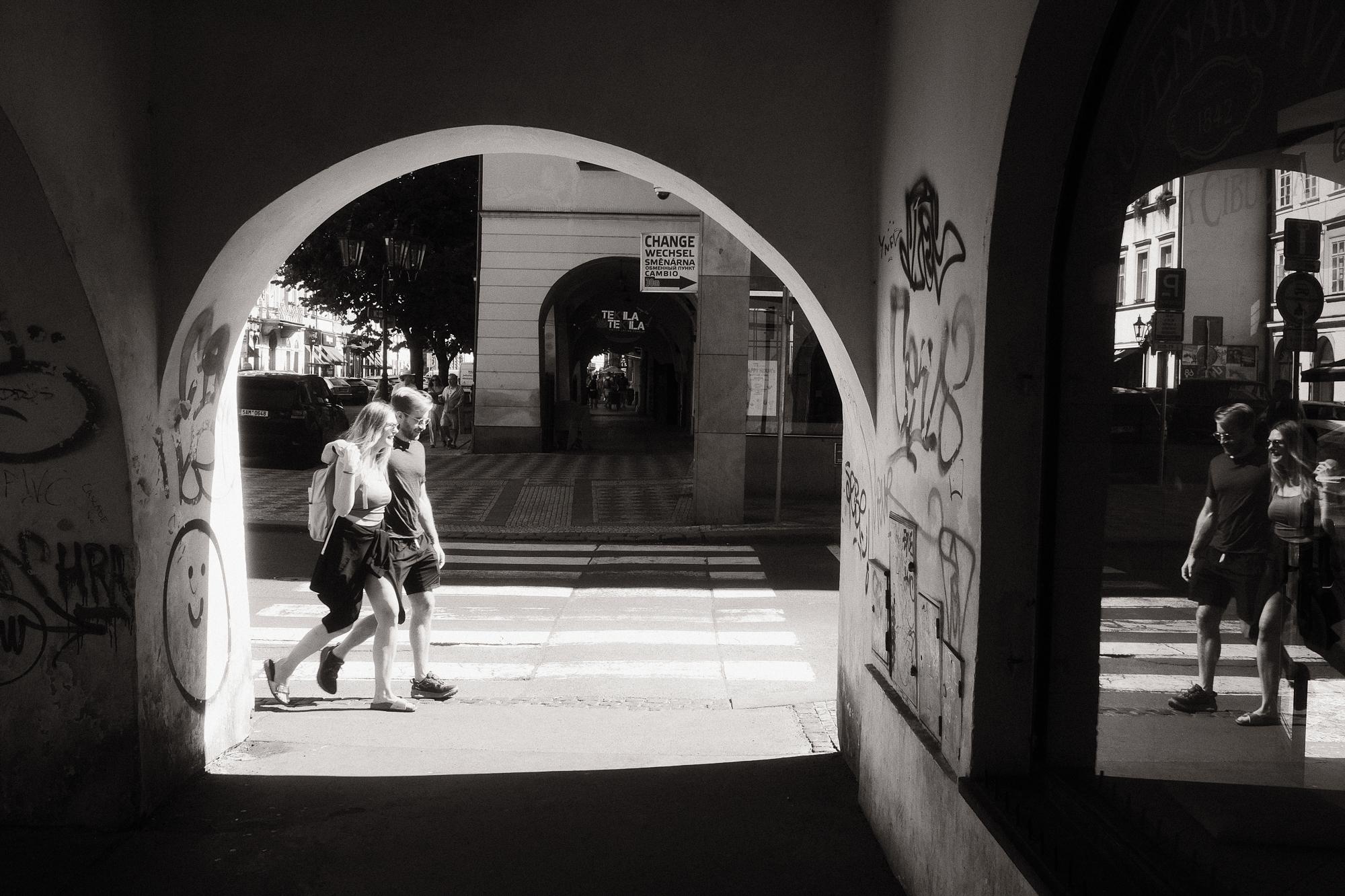 Čiernobiela fotografia dvojice prechádzajúcej cez priechod pre chodcov pod oblúkovým podjazdom.