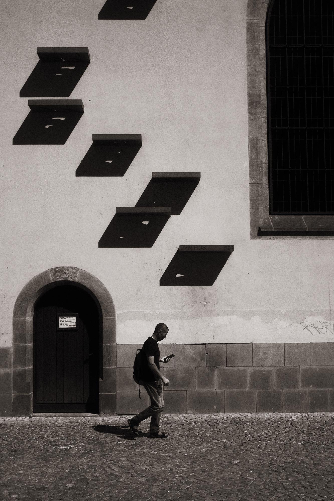 Čiernobiela fotografia zachytáva osobu, ktorá kráča po kamennej dlážke pri starom múre s oblúkovitými dverami a výraznými tienmi na stene.