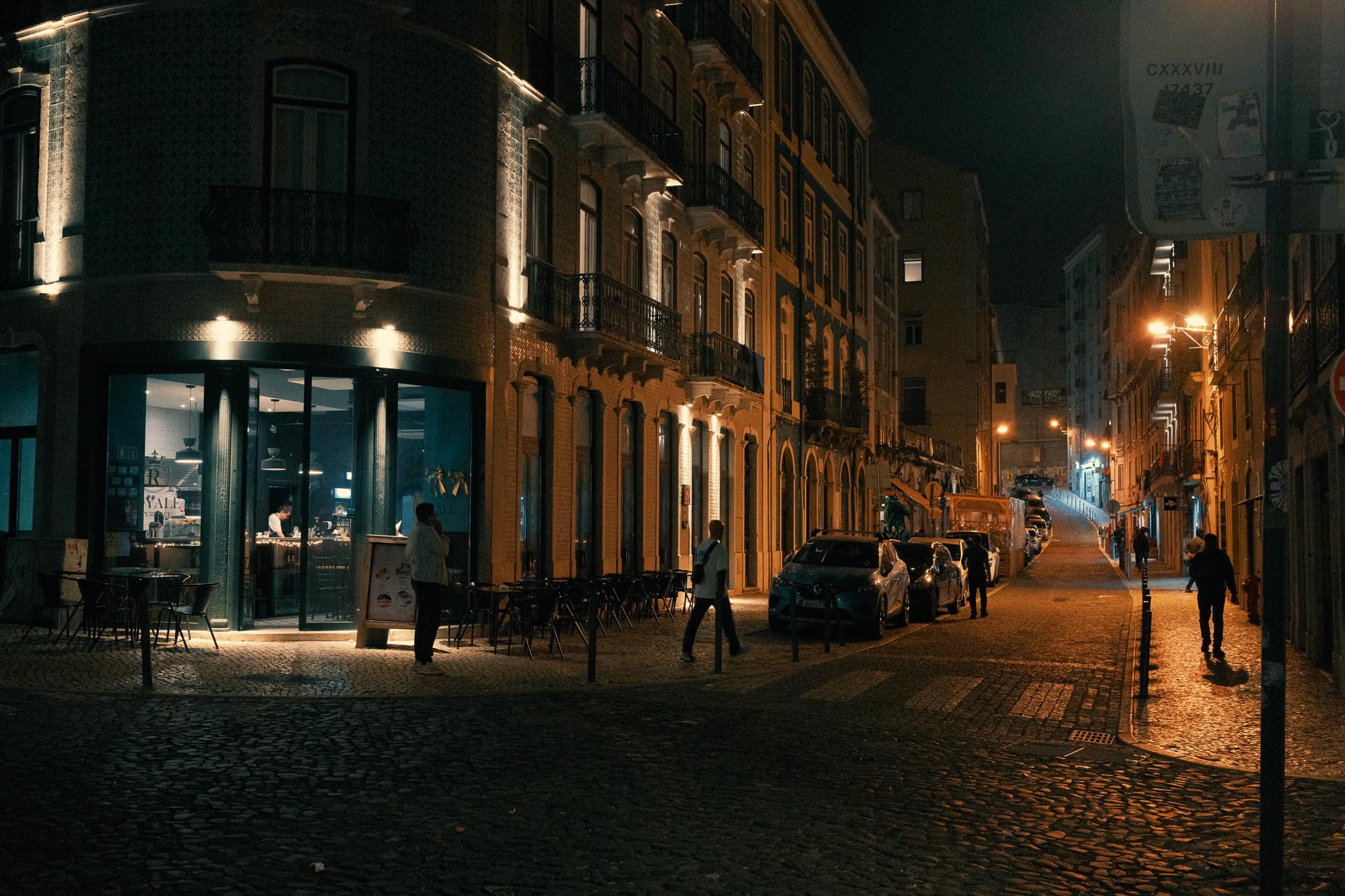 Nočná ulica s osvetlenými budovami, bistro s veľkými oknami na rohu. Kontrast studeného a teplého svetla.