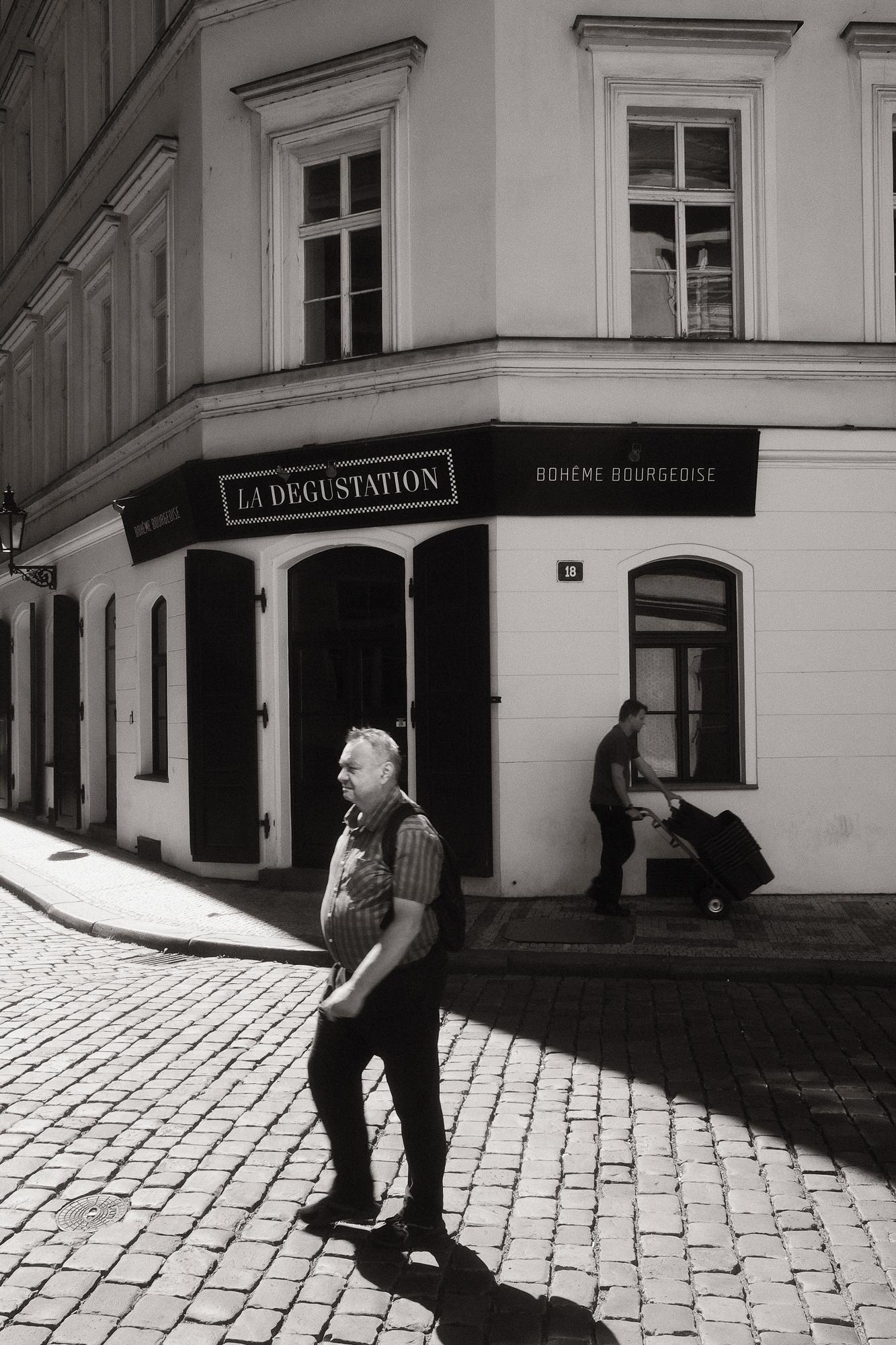 Čiernobiela fotografia muža kráčajúceho po ulici v silno kontrastnom slnečnom svetle.