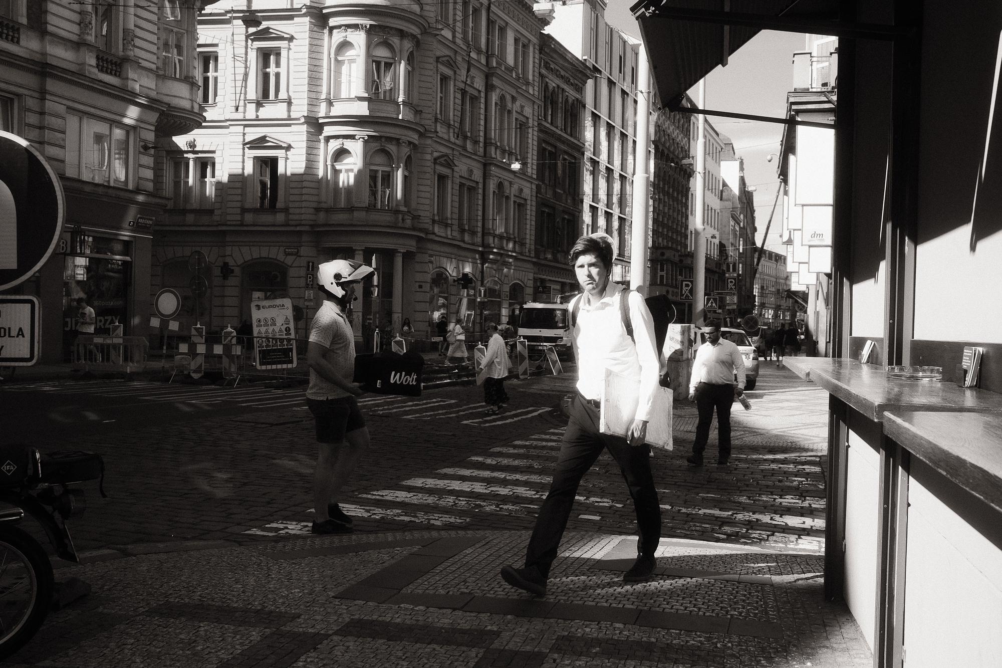 Čiernobiela fotografia rušnej mestskej ulice so starými budovami v pozadí. V popredí človek v bielej košeľi a aktovkou prechádzajúci po prechode.