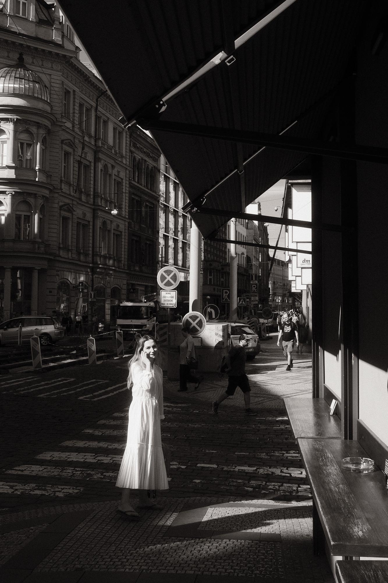 Čiernobiela fotografia pražskej ulice. V popredí sa nachádza telefonujúca žena v bielych šatách prechádzajúca cez pás slnečného svetla.
