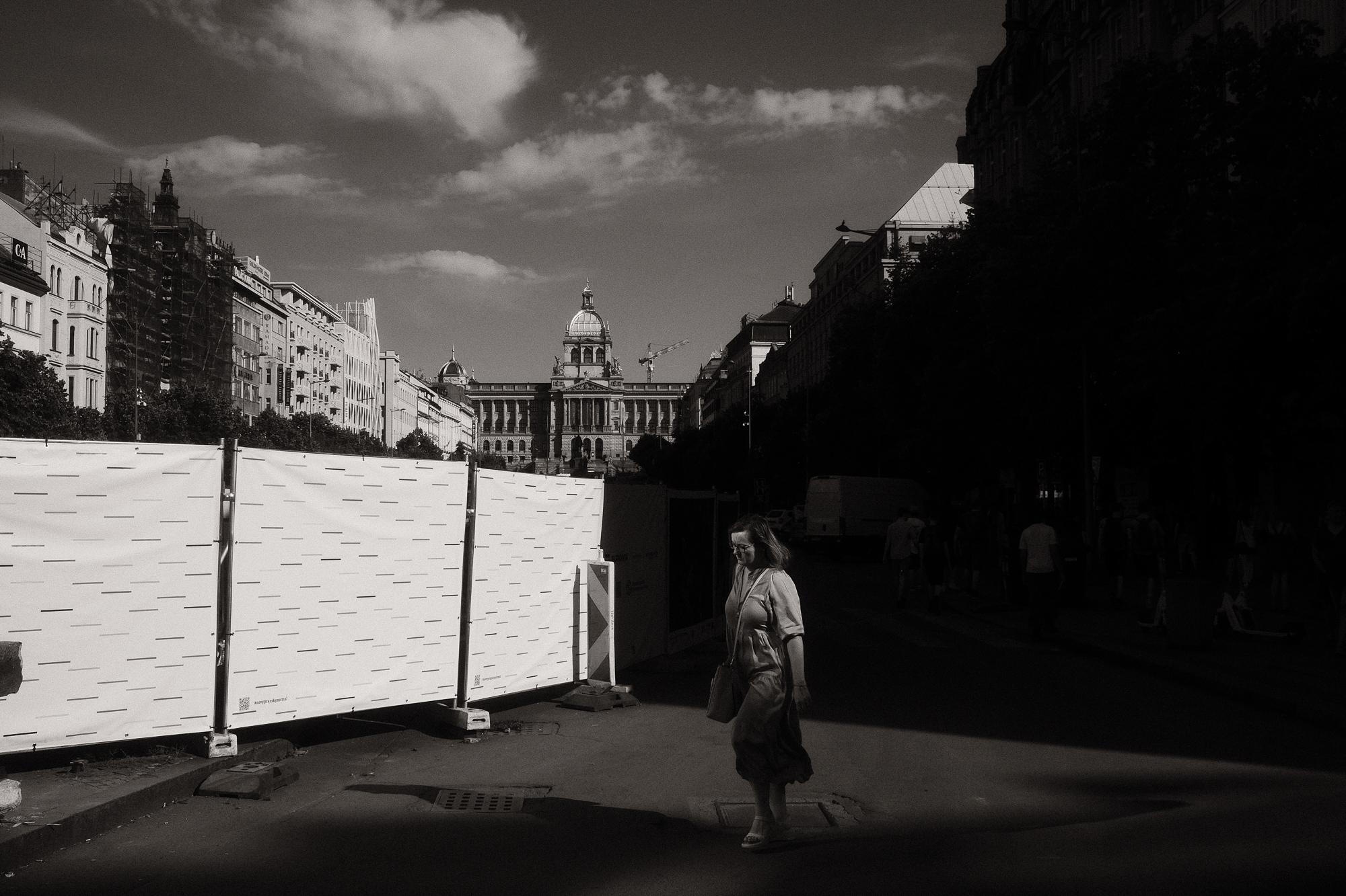 Čiernobiela fotografia Václavského námestia s historickou budovou Muzea v pozadí. Žena prechádza popri stavenisku s bielou ohradou, obklopená budovami a slnečným svetlom. Praha.