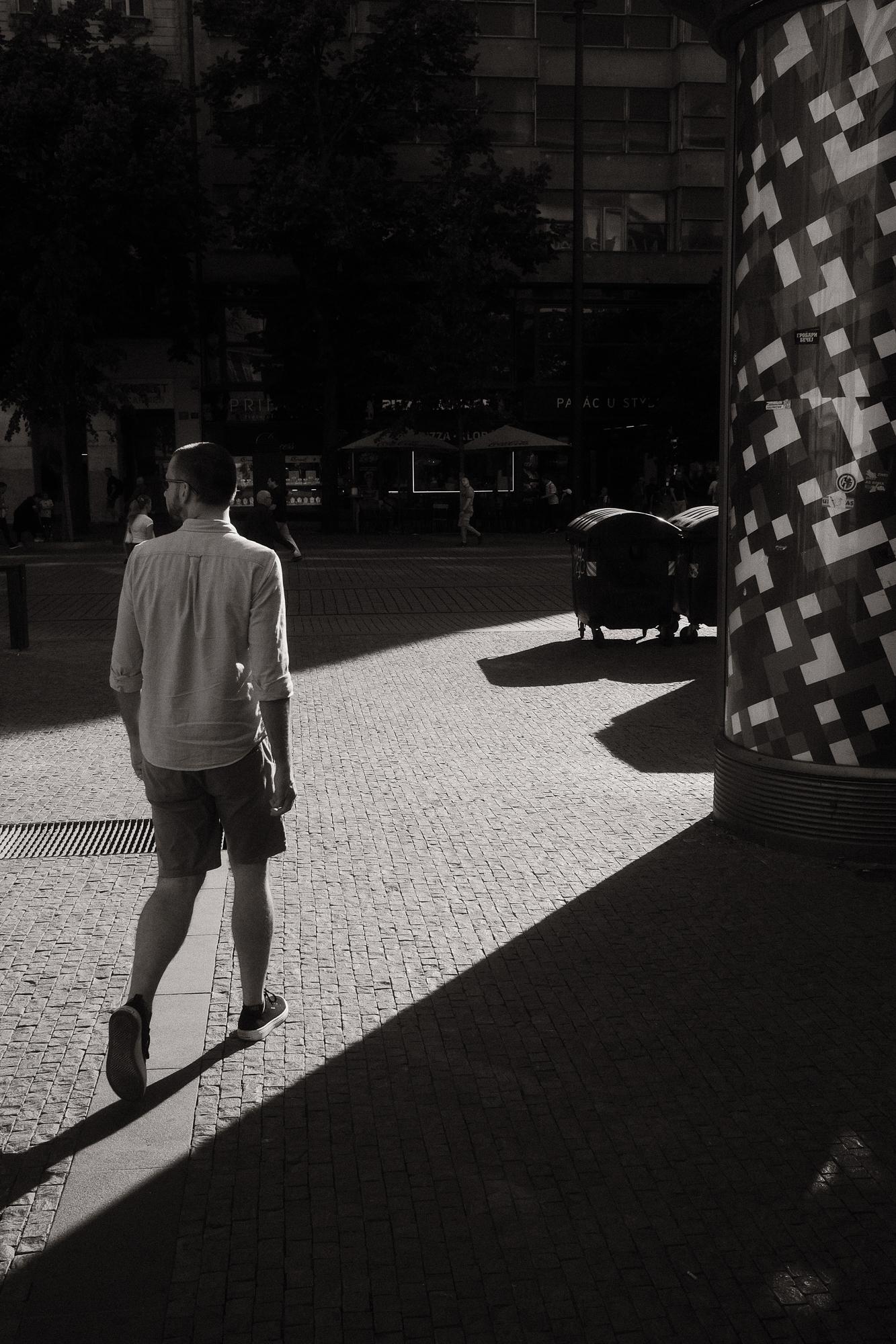 Čiernobiela fotografia muža kráčajúceho po dlažbe, s dlhým tieňom padajúcim na zem.