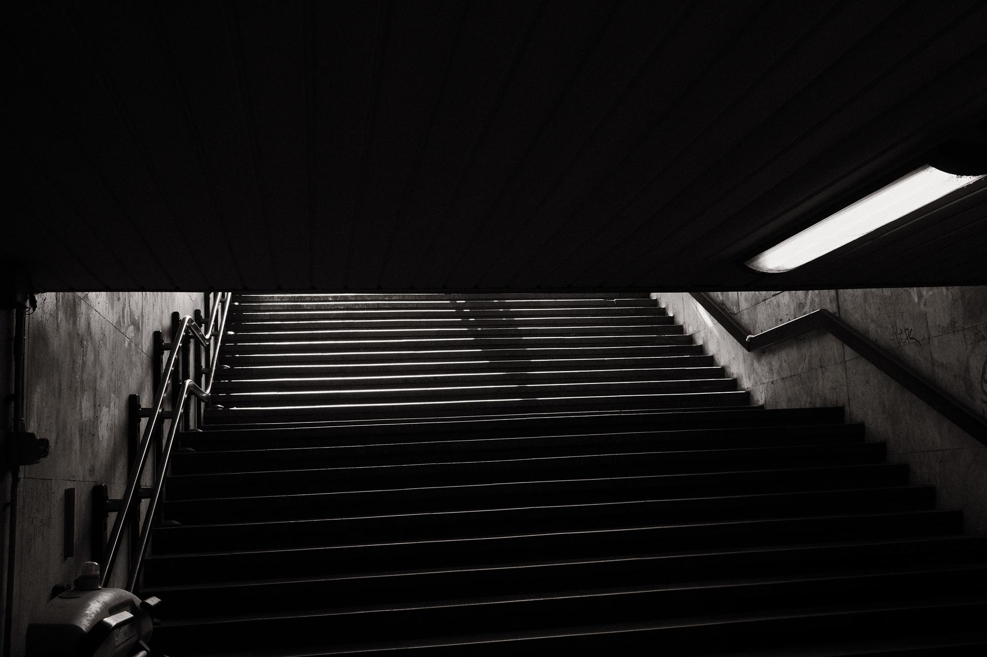 Čiernobiela fotografia zachytáva podchod so schodami vedúcimi smerom nahor, zhora vidíme tieň prichádzajúcej postavy.
