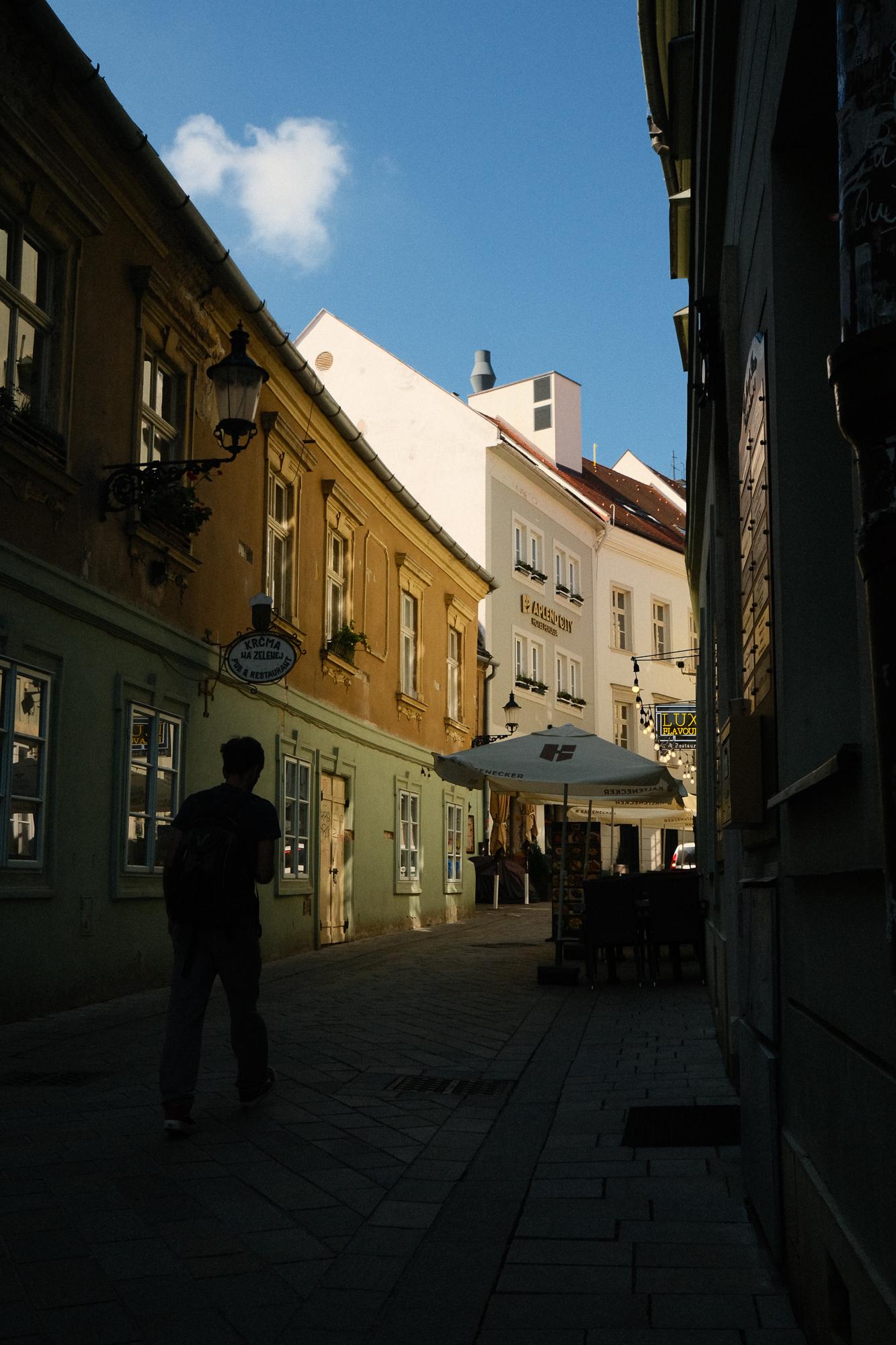 Úzka ulička v Bratislave s farebnými historickými budovami osvetlenými slnečným svetlom a modrou oblohou v pozadí.