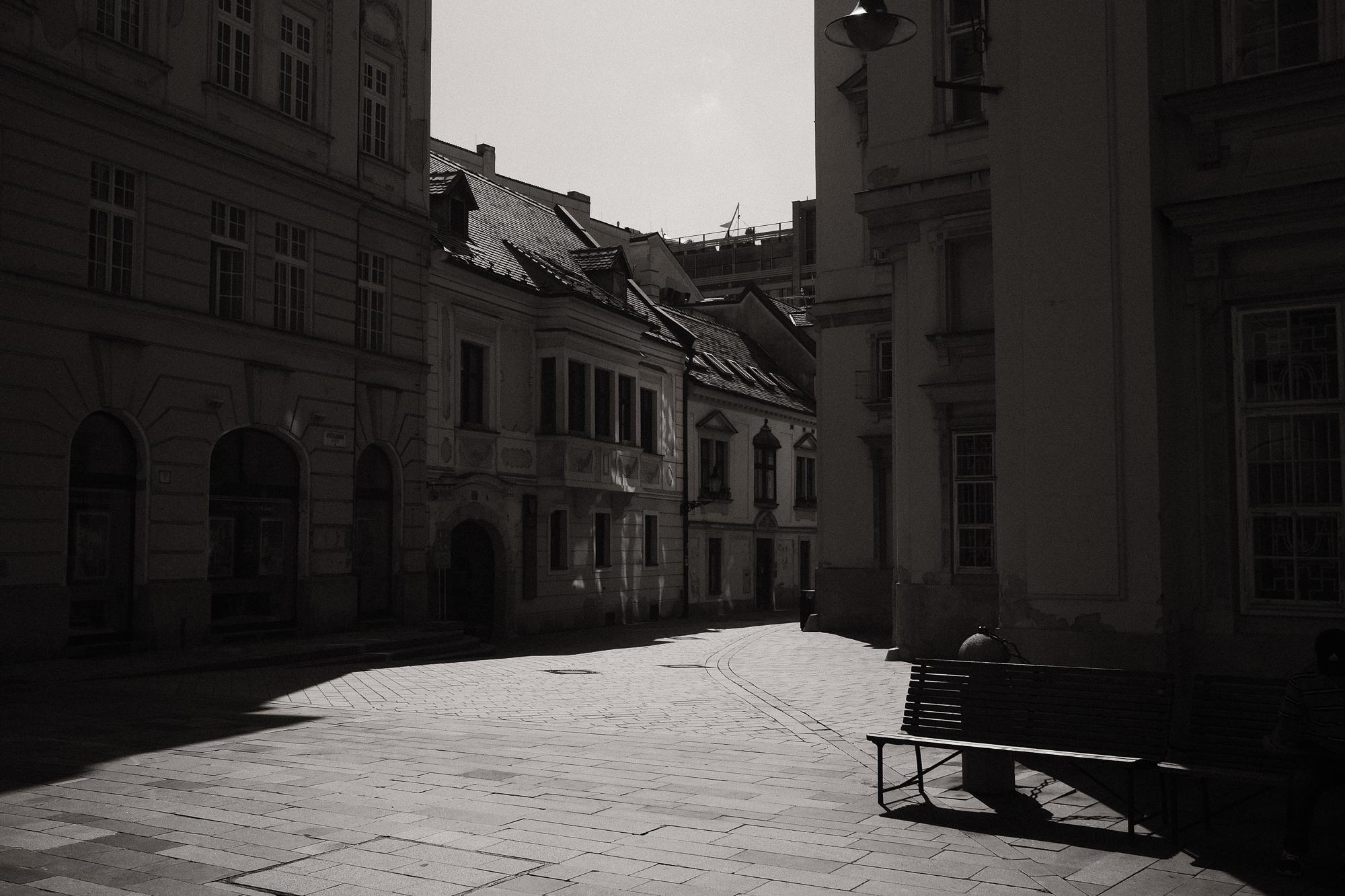 Čiernobiela fotografia mestského námestia s historickými budovami, osvetleného pruhmi slnečného svetla. Bratislava.