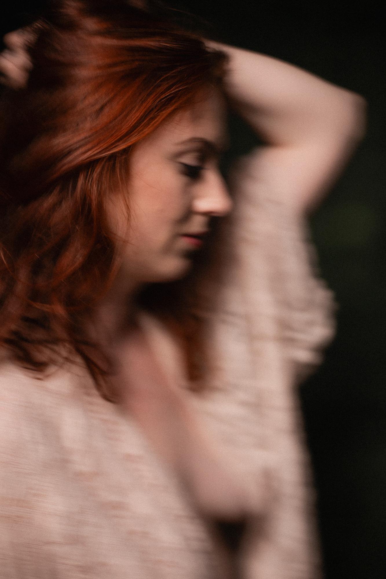 Rozmazaný portrét ženy s hrdzavými vlasmi, ktorá má zatvorené oči a ruku položenú na vlasoch na tmavozelenom pozadí.