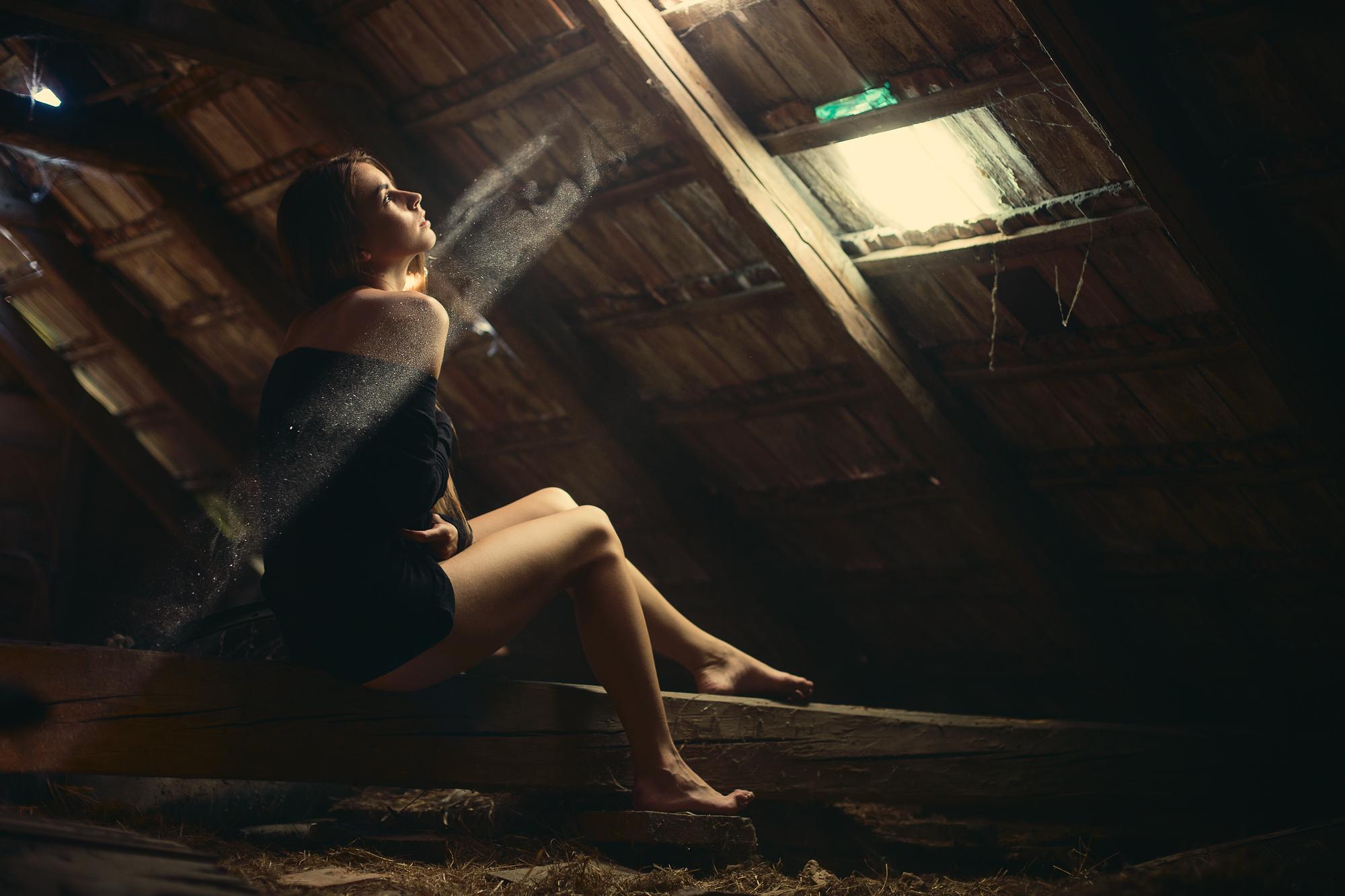 Žena sediaca na tráme v podkroví, oblečená v čiernych šatách, osvetlená lúčom svetla viditeľnom v čiastočkách prachu.