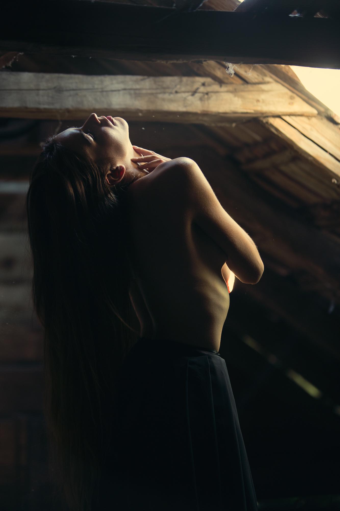 Portrét ženy s odhalenými ramenami a hlavou zaklonenou dozadu, osvetlenou jemným svetlom prenikajúcim cez strešný otvor v tmavej drevenej povale.