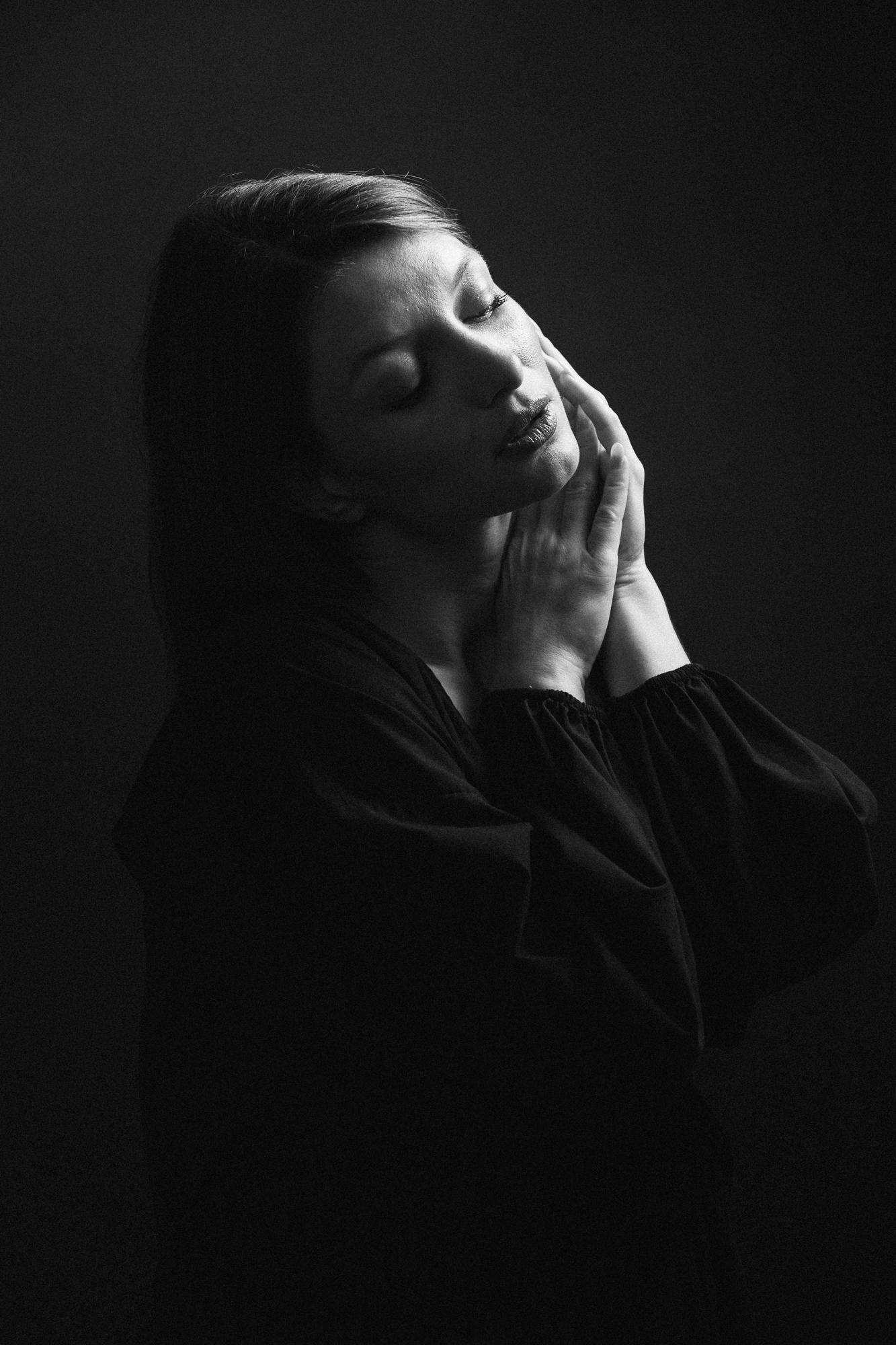 Čiernobiela portrétna fotografia ženy so privretými očami v jemnom osvetlení, jemne si opierajúca tvár o ruky.