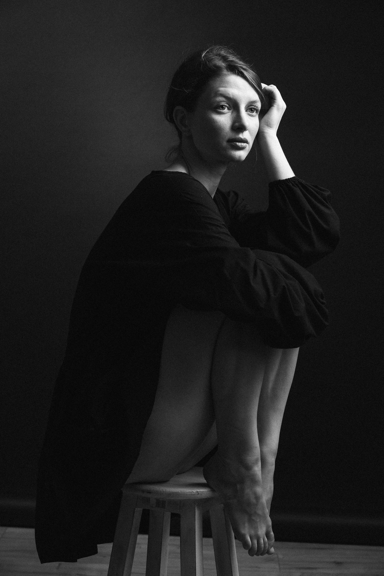 Black and white portrait of a woman sitting on a wooden stool in a loose black dress, knees drawn up and feet bare, one hand in her hair, against a dark background. Studio Pezinok.