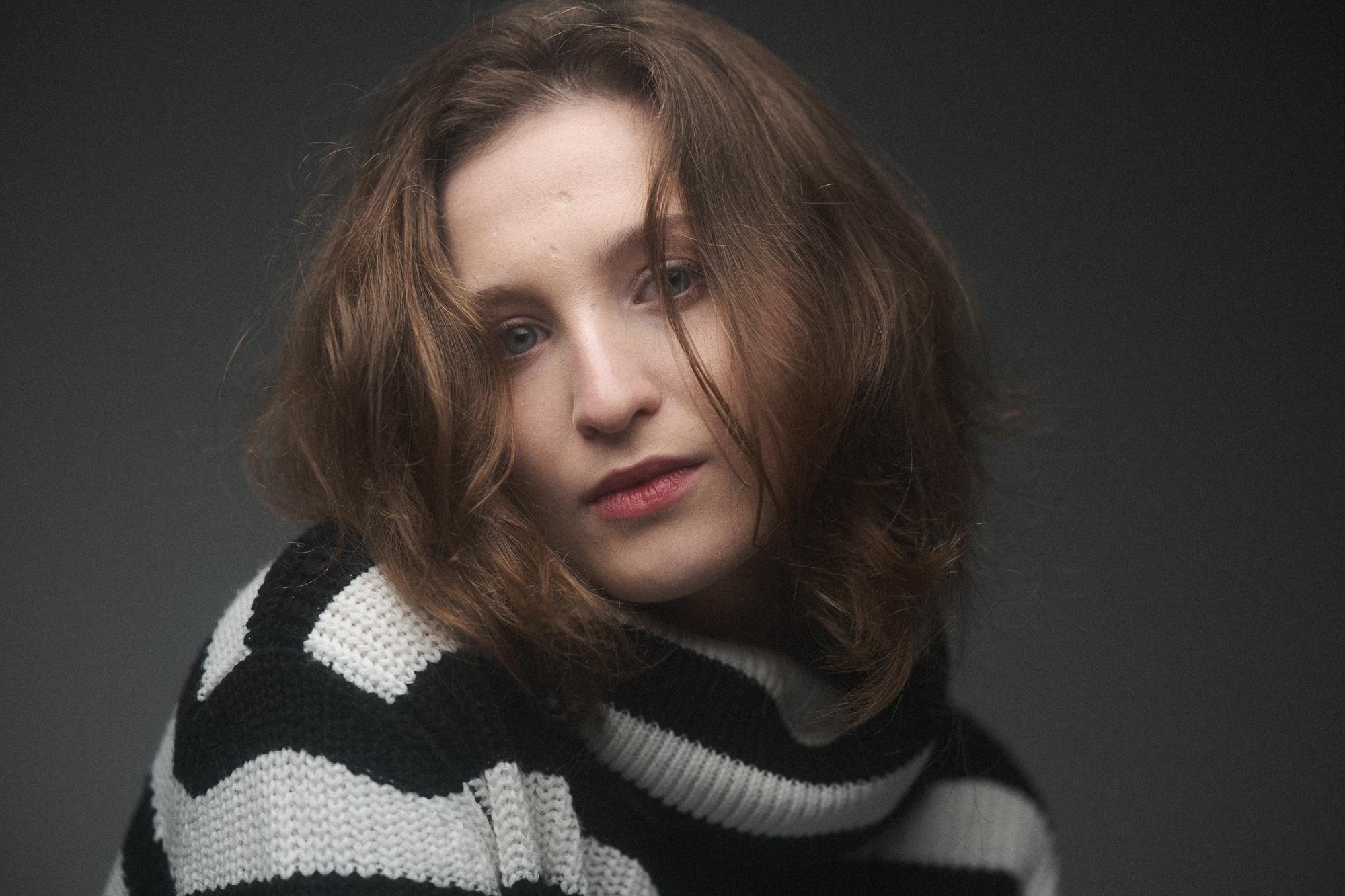 Portrét ženy s hnedými vlasmi v čiernom a bielom pruhovanom svetri, s upreným výrazom tváre, na sivom pozadí.