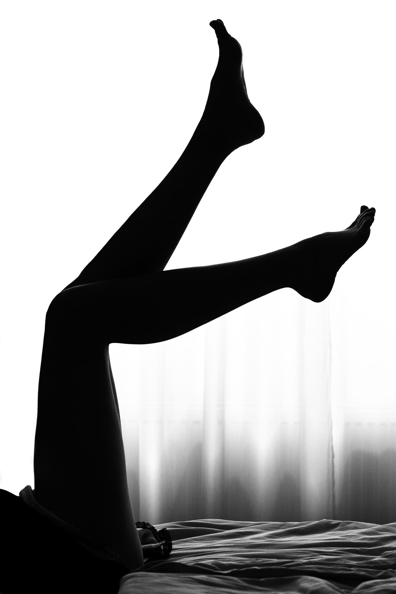 Black and white artistic nude photograph capturing the silhouette of female legs pointing upwards from bed, in front of sheer curtains. Prague.