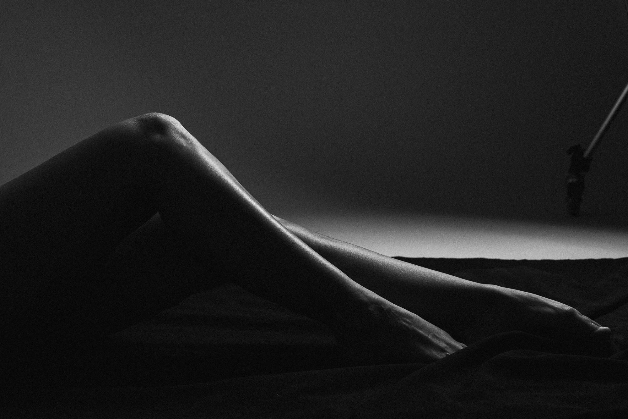 Black and white artistic nude photograph featuring the graceful lines of legs highlighted by dramatic lighting against a dark background. Studio Pezinok.