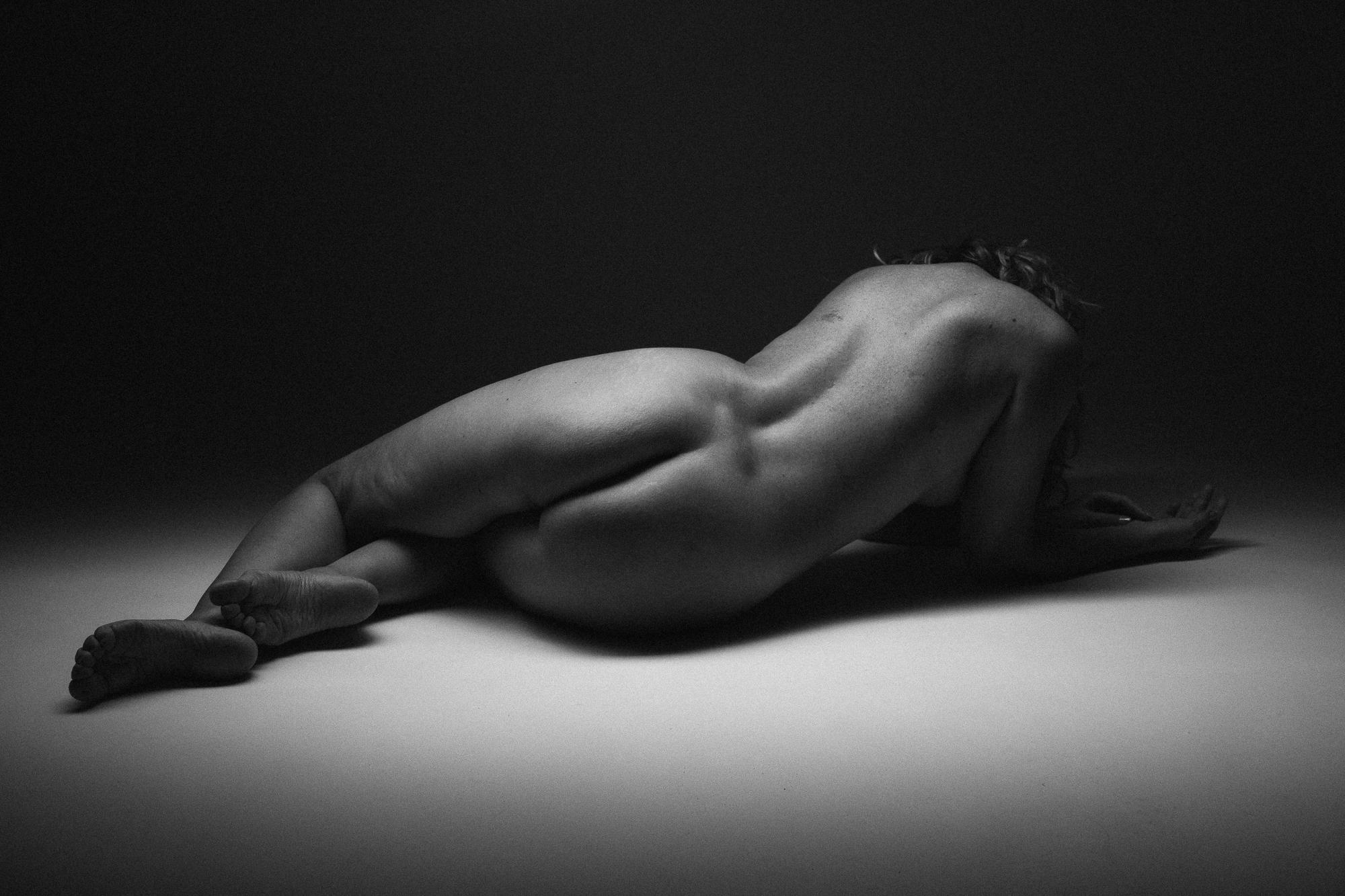 Black and white artistic nude photograph depicting a nude figure lying on the side in dramatic lighting. Studio Pezinok.