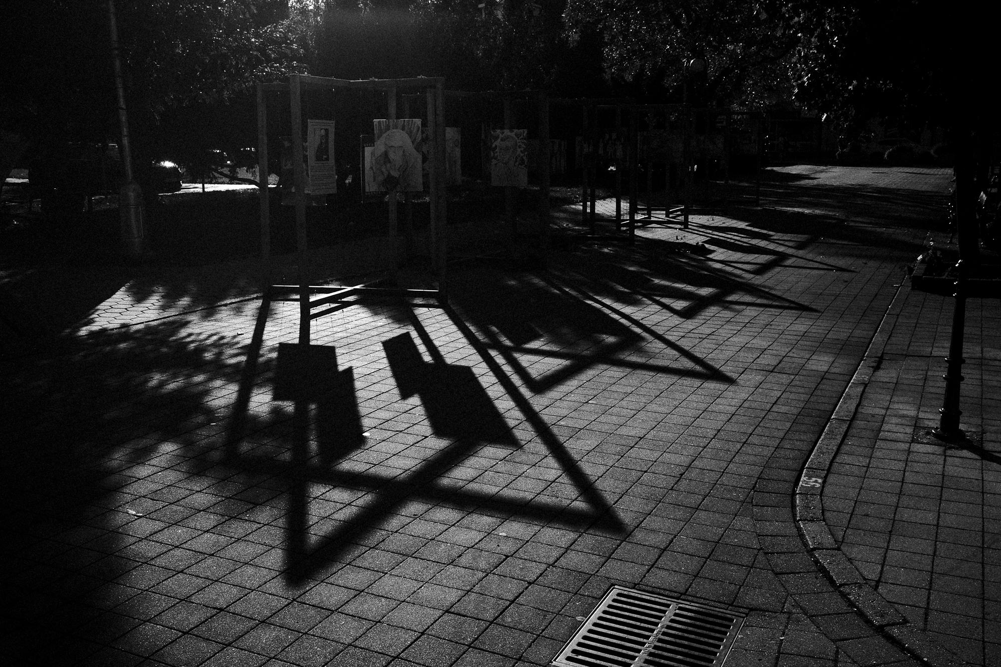 Čiernobiela fotografia mestskej ulice s tmavými tieňmi padajúcimi na dlažobné kocky. Na pozadí sú viditeľné rámy obrázkov a stromy vrhajúce tieň.