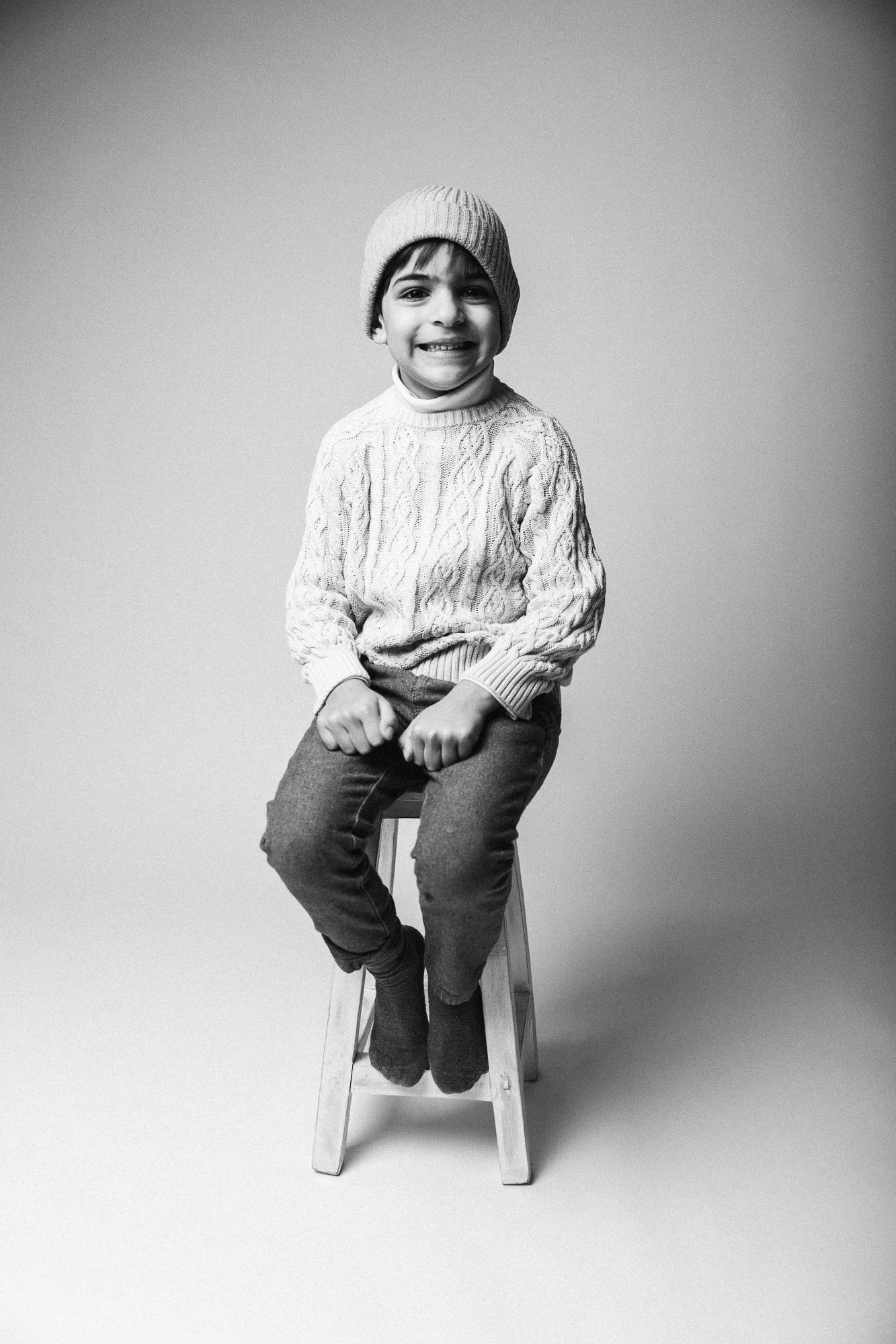 Čiernobiely portrét malého chlapca sediaceho na stoličke, oblečeného v pletenom svetri a čapici, s úsmevom na tvári. Ateliér Pezinok pri Bratislave.