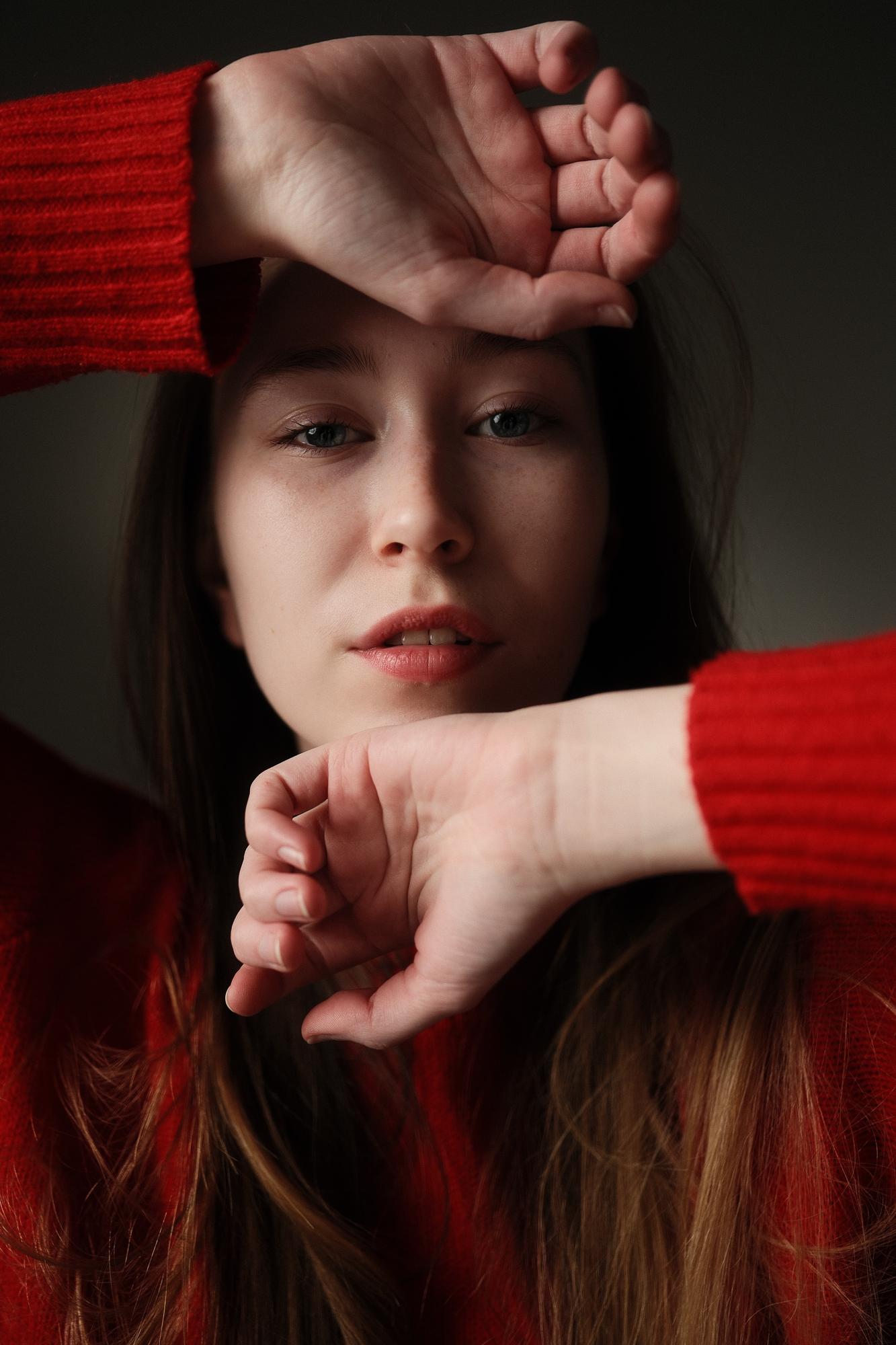 Portrét mladej ženy v červenom svetri, tvár orámovaná zdvihnutými rukami, jemne osvetlená na tmavom, tlmenom pozadí. Ateliér Pezinok