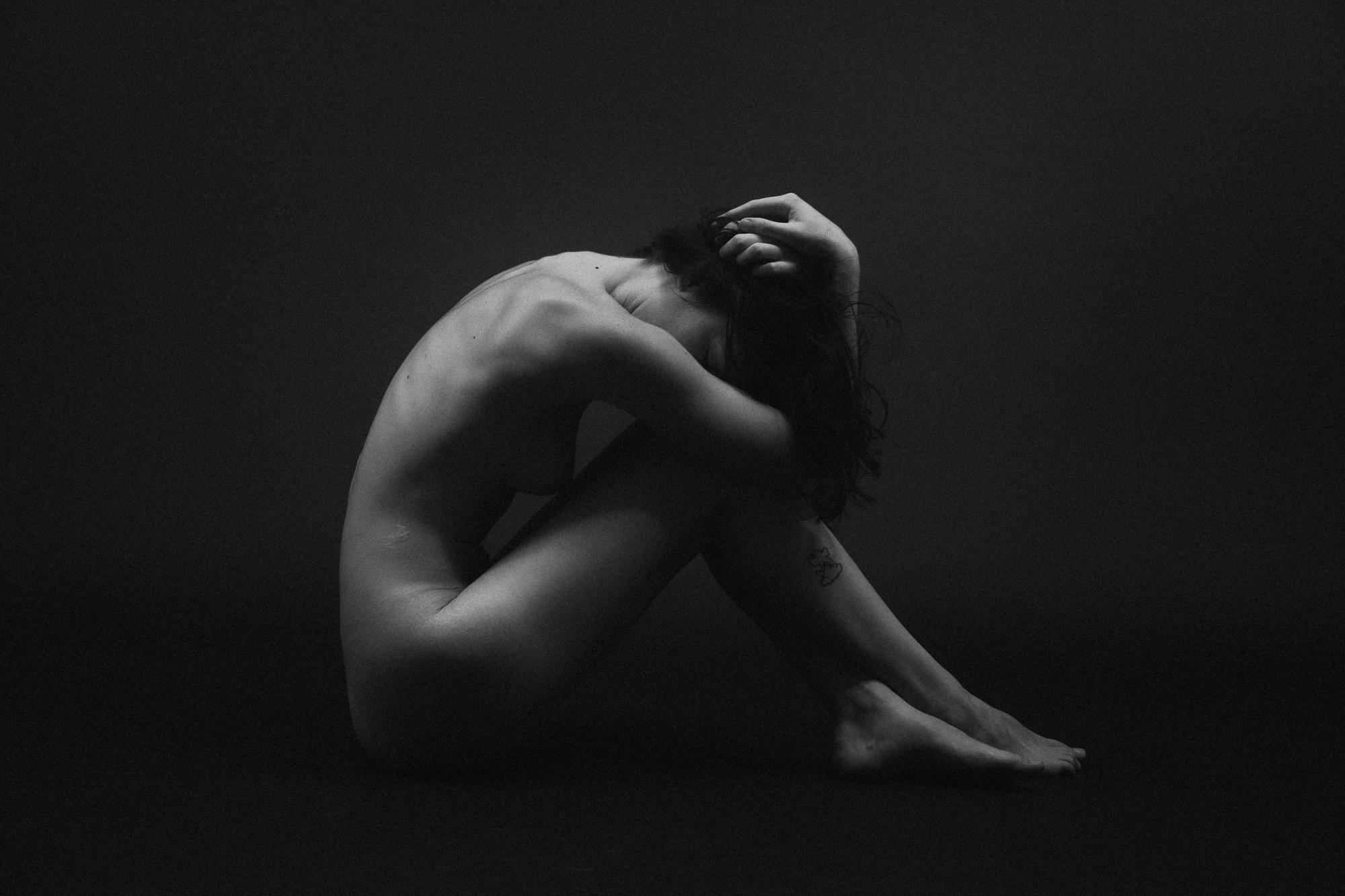 Black and white artistic nude photograph. Young woman sits on the floor, subtly covered by her own arms and hair, in an expressive pose. Studio Pezinok.