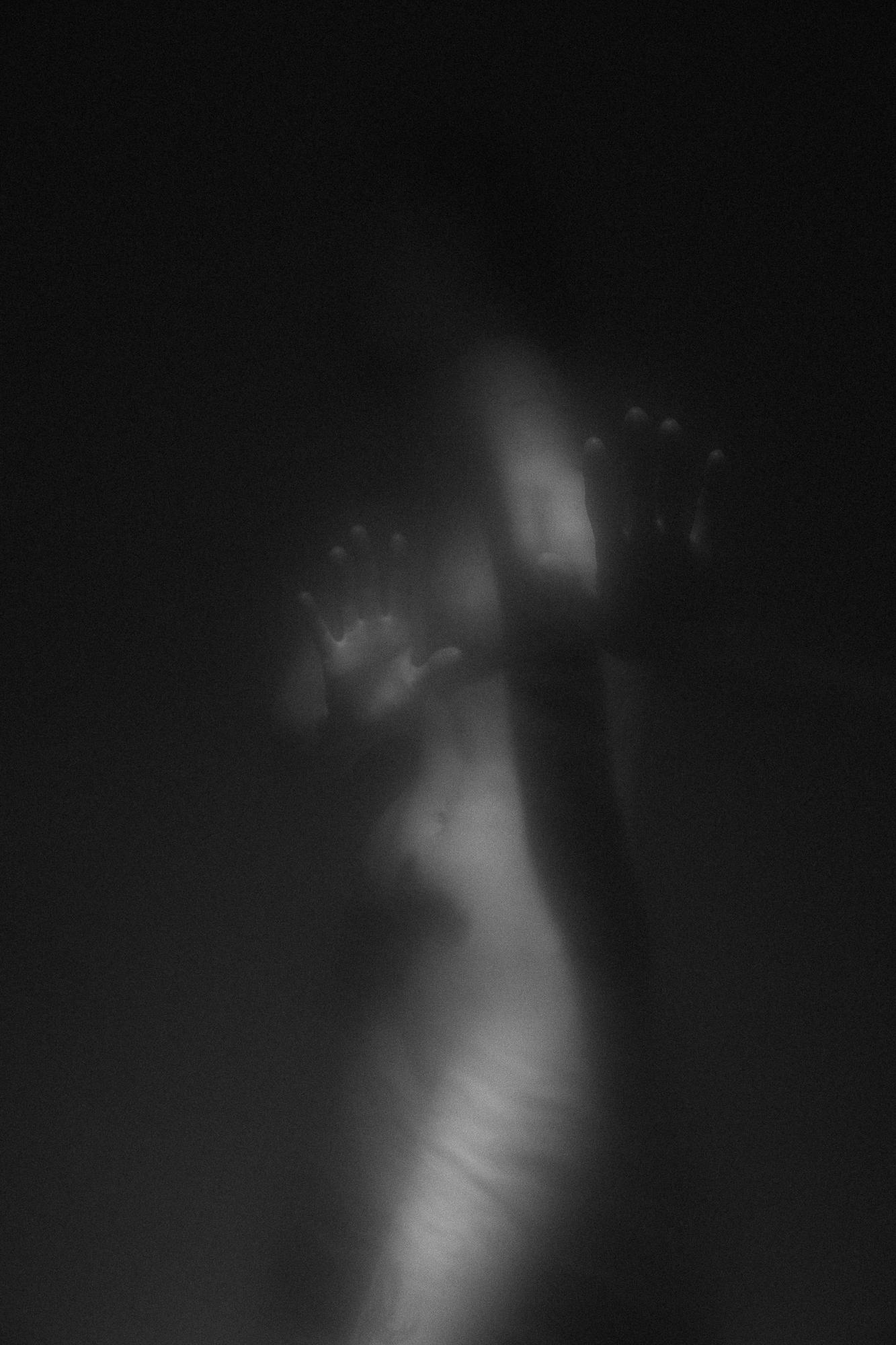 Black and white artistic nude photograph depicting a blurred figure with raised hands, creating a sense of soft light and shadows. Studio Pezinok.