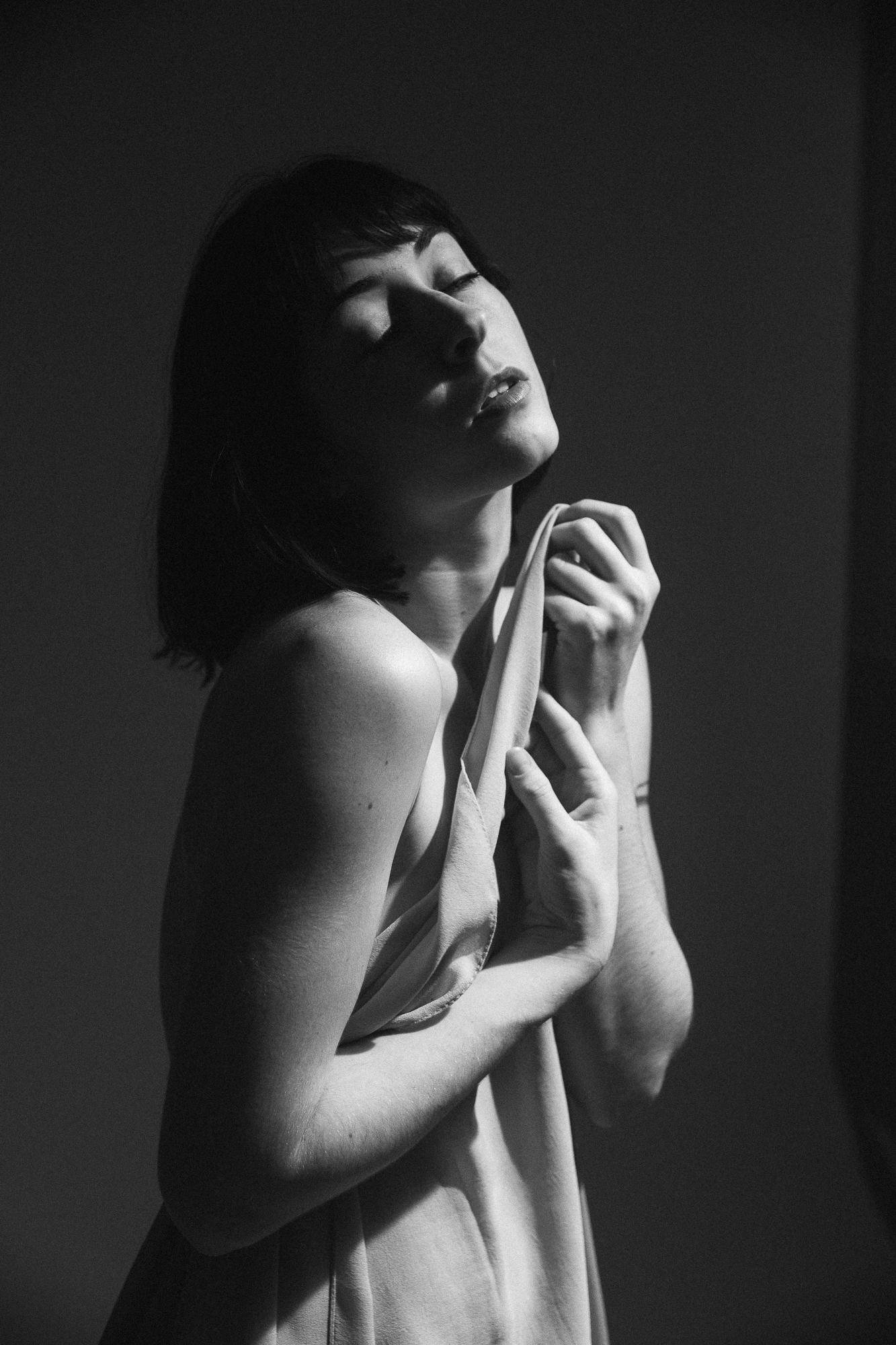 Čiernobiely portrét ženy zakrývajúcej sa látkou pri bočnom jemnom svetle.