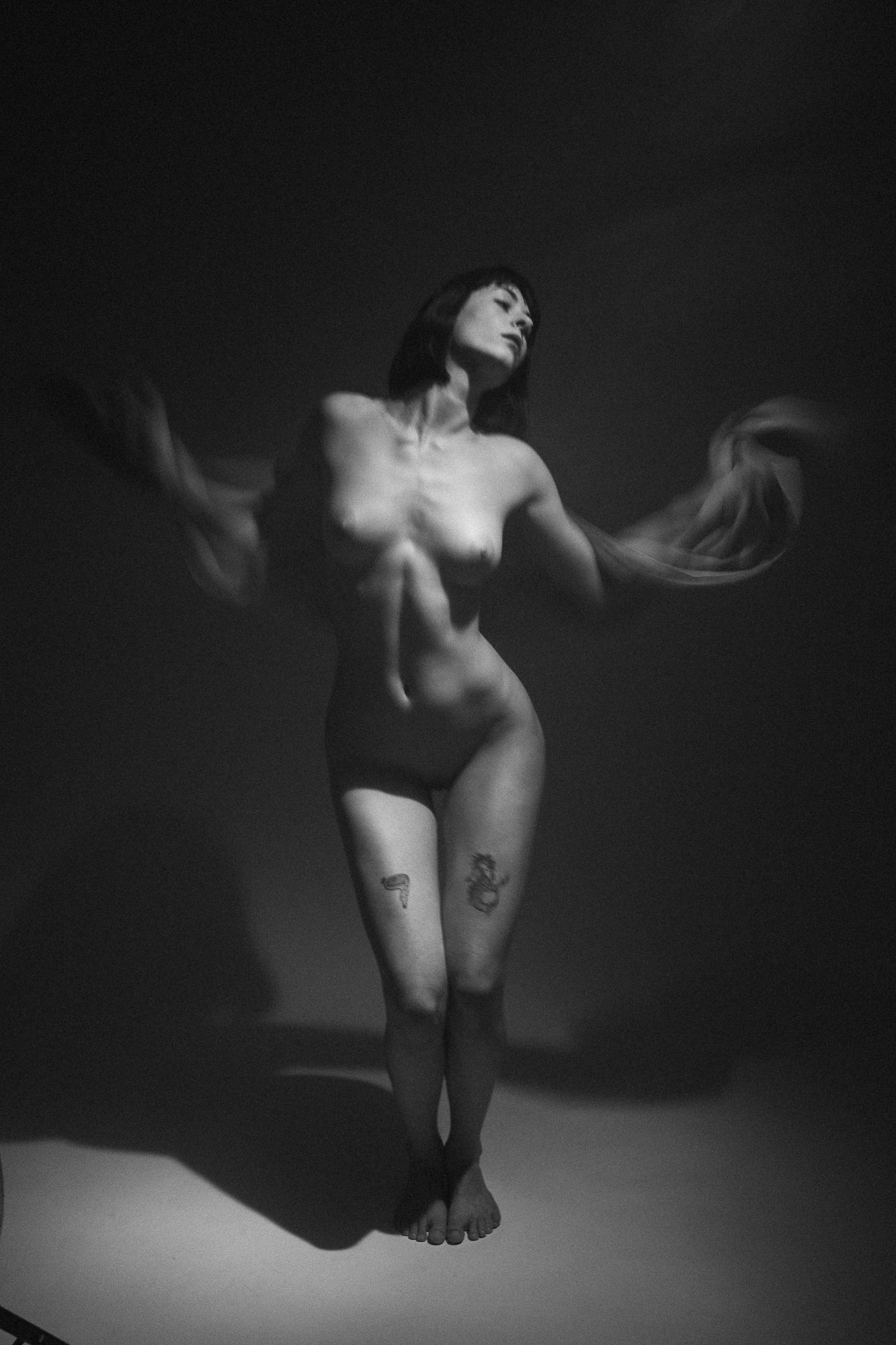 An intimate black and white artistic nude photograph depicting a woman's figure in motion emphasized by a blurred cloth in her hands. Studio Pezinok.