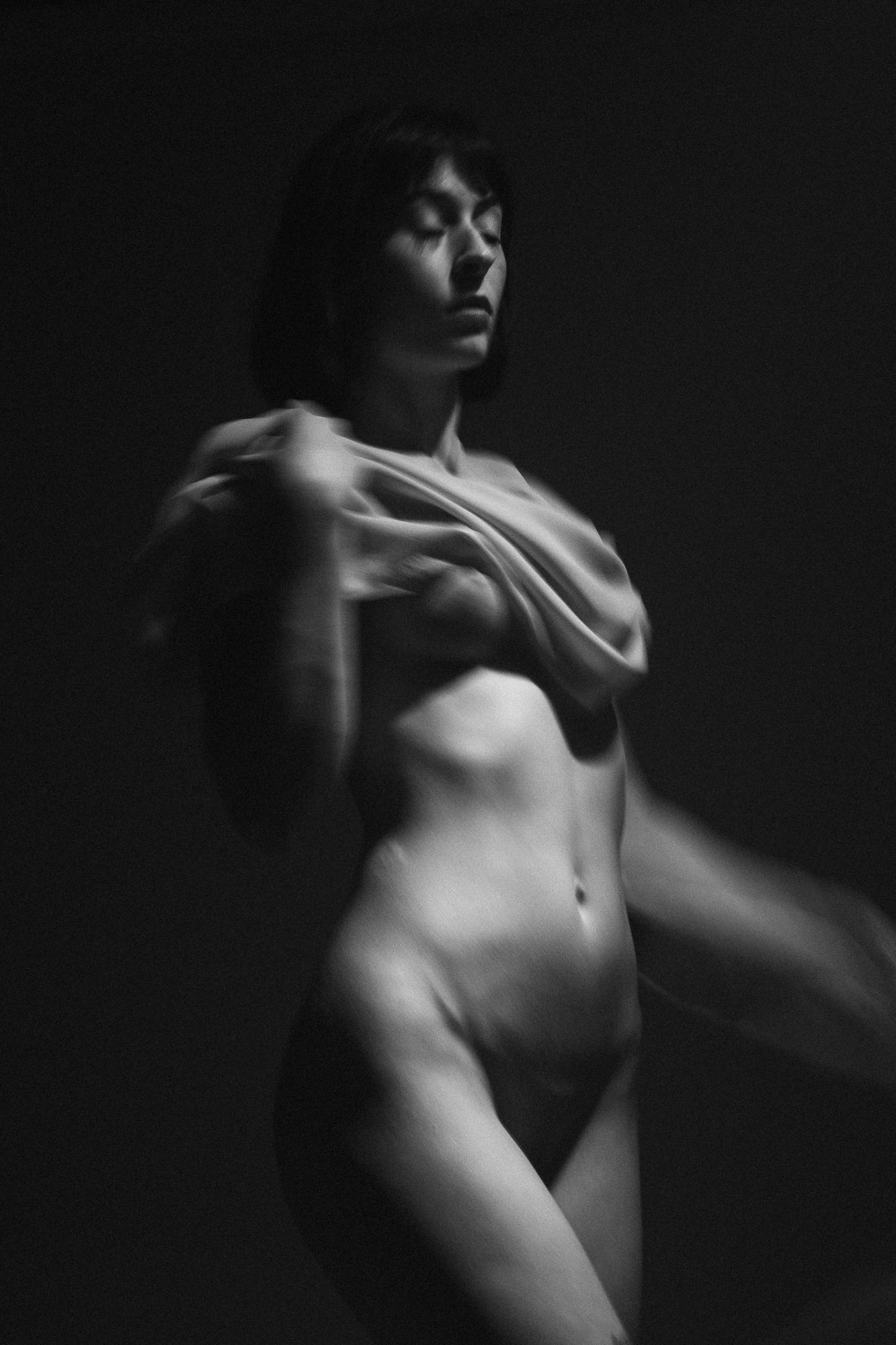 Black and white artistic nude photograph depicting a female figure, softly lit, in an artistic movement with fabric. Studio Pezinok.