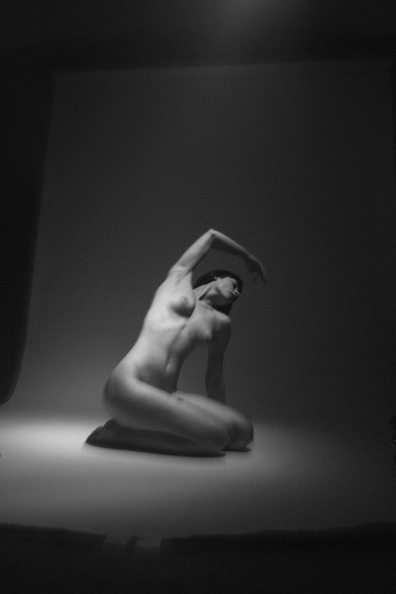 Black and white artistic nude photograph captures a figure seated on the floor with head tilted back and one arm raised. The image exudes elegance and delicacy. Studio Pezinok.